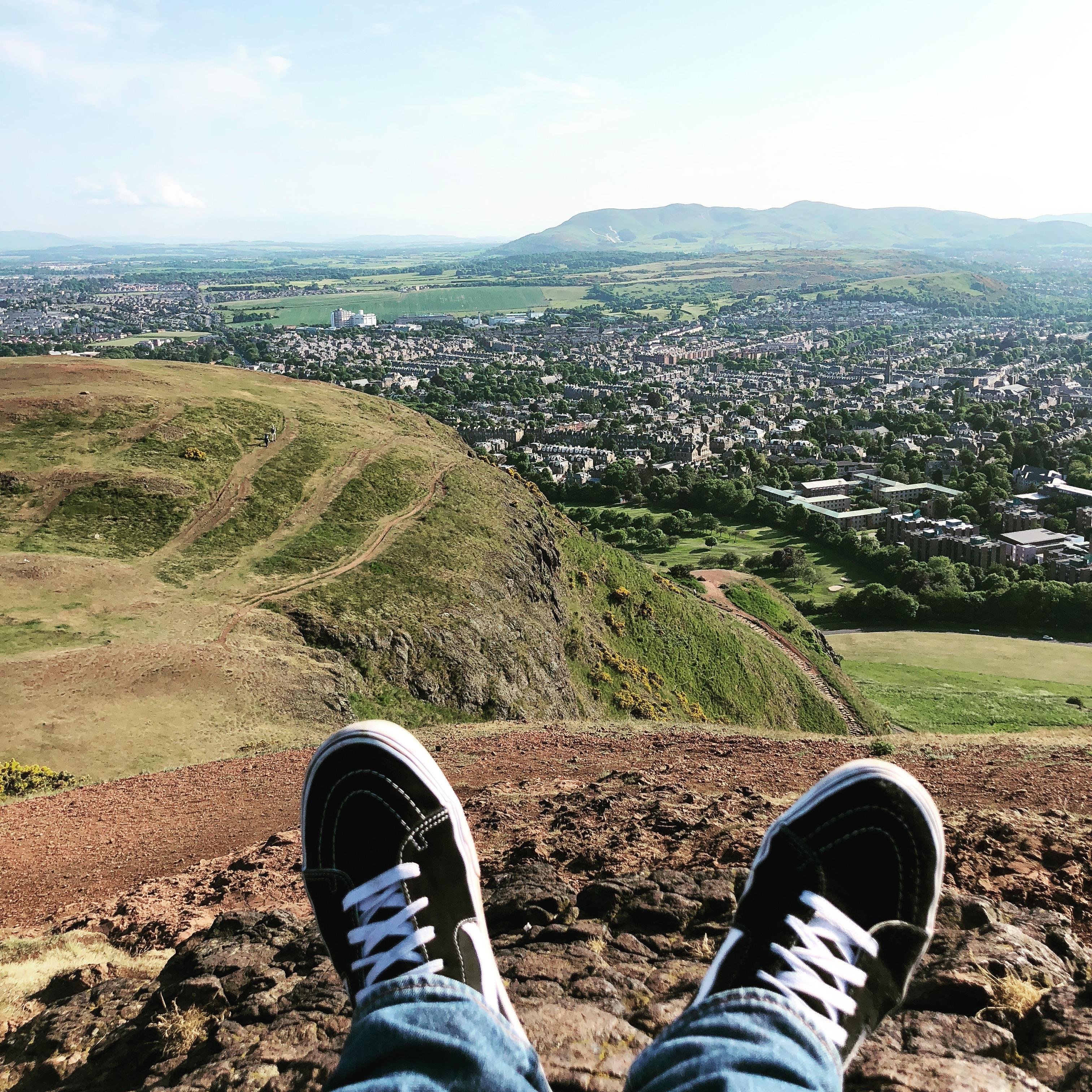The image shows a beautiful panoramic view from a high vantage point over a lush, green landscape and a town. In the foreground, a person's legs are extended with their feet wearing sneakers, dangling over a rocky edge, suggesting they are sitting and enjoying the view from a high location. The visible paths across the greenery indicate popular hiking or walking trails. In the distance, there is a town spread out with various buildings, and beyond the town, a range of hills or mountains can be seen under a clear sky. This setting might perhaps be somewhere known for its scenic hikes and views.