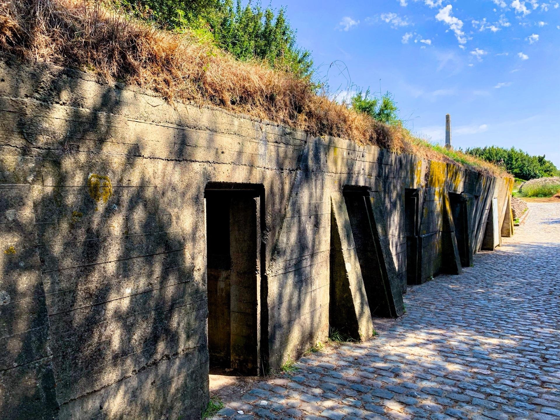 This image shows a series of old, weathered concrete bunkers with multiple entries aligned in a row. These are typically underground shelters partially built into a grassy mound, designed to protect from blasts and projectiles. The bunkers feature open doorways and buttresses between them for structural support. A cobblestone pathway runs in front of the entrances, indicating that this is likely a historical or military site being preserved. The condition and design suggest they might be from the World War I or II era.