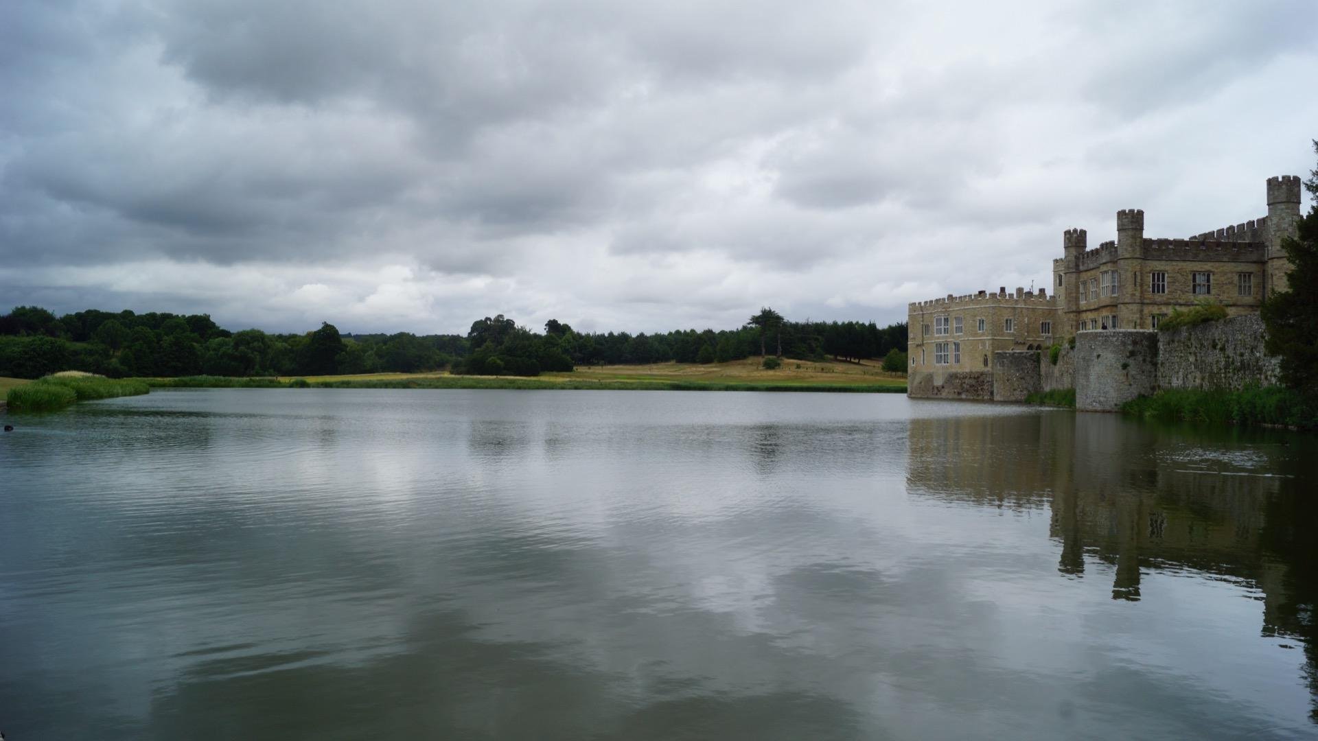 The image features a scenic view of a large body of water with a historic-looking castle on its edge. The castle has an old, robust architecture typical of medieval or early modern European structures, presented with stone walls and what appear to be defensive features. The sky is overcast, suggesting a cloudy day, and there is lush greenery in the background across the lake, enhancing the peaceful yet dramatic atmosphere of the setting. This looks like it could be a location in Europe, possibly the UK given the style of the castle and the landscape.