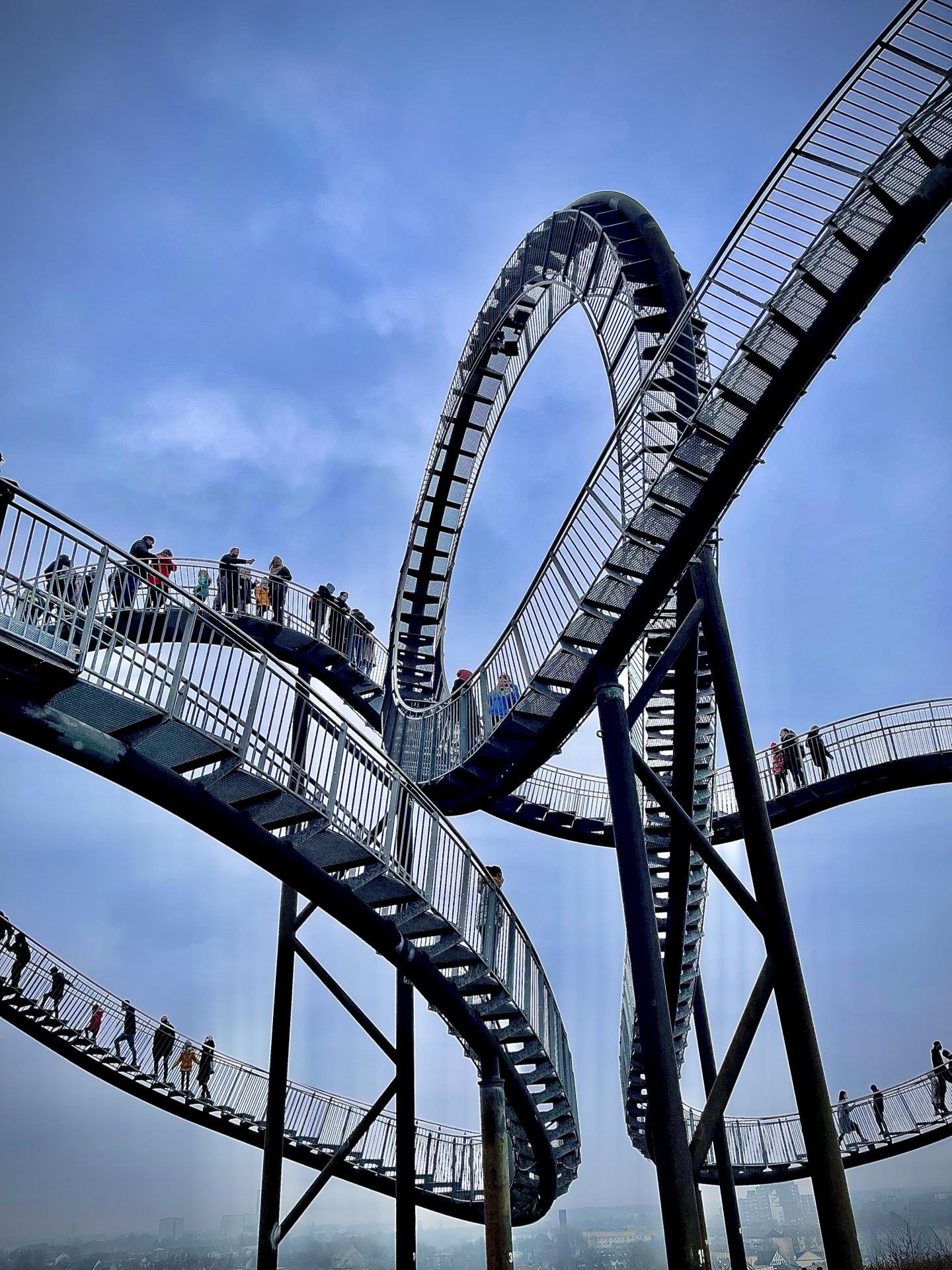 Outdoor spiral staircase sculpture with a loop at the top, constructed of metal with mesh railings, against a hazy sky. People are ascending and standing on various parts of the walkways, observing the surroundings.
