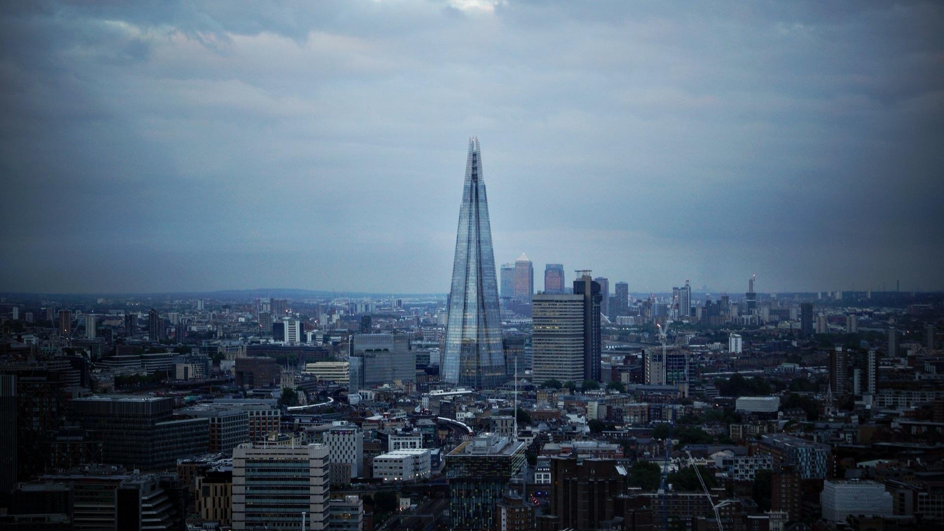 This image features a panoramic view of London's skyline at a time when the sky looks overcast. Dominating the center of the image is The Shard, which is a notable skyscraper in London known for its distinctive, sharply pointed shape. Surrounding The Shard, you can see a spread of other buildings that vary in height and design, typical of a bustling city landscape. The overall mood conveyed by the cloudy sky and distant cityscape suggests a vast, gently bustling urban environment.