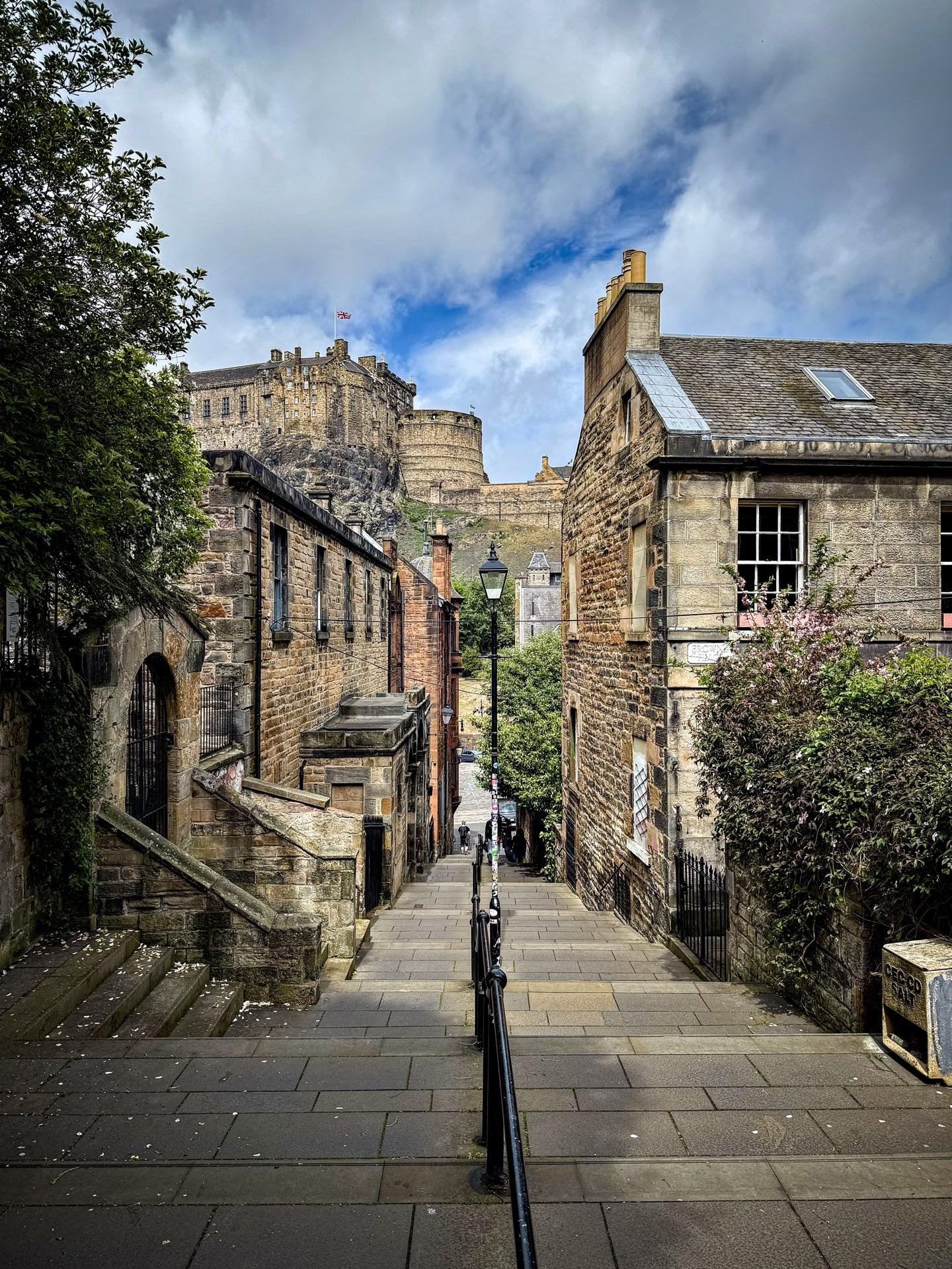 Narrow cobblestone street leading up to a grand, ancient stone castle with flags flying on its battlements, flanked by traditional stone buildings with a blue sky and scattered clouds above. A metal railing runs along the left side of the street, guiding the path of pedestrians walking along the pavement.