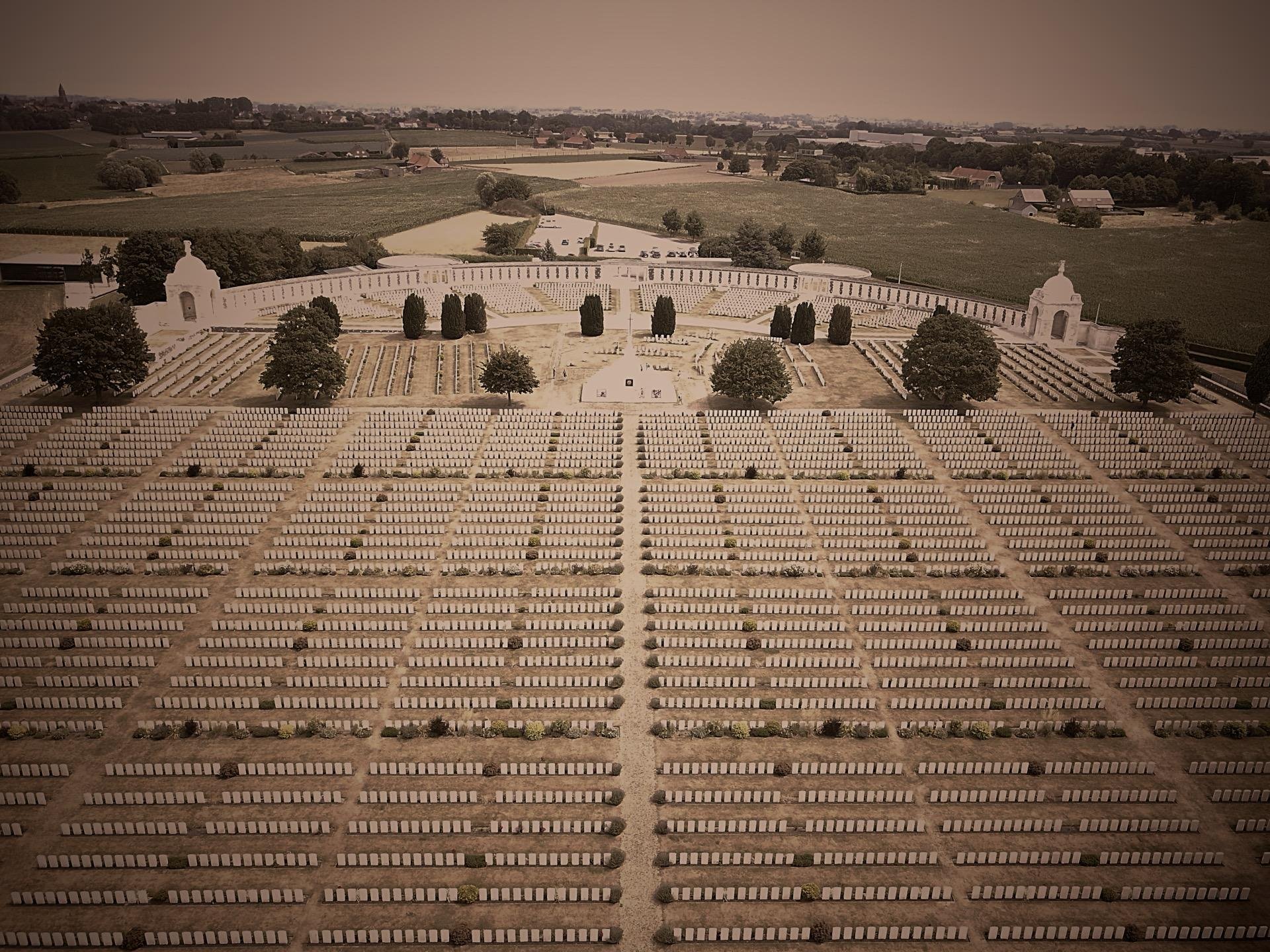This image shows a large military cemetery, evident from the orderly rows of grave markers. In the center, there appears to be a monument or a memorial chapel, surrounded by landscaped grounds including paths and trees. The cemetery is expansive, with a vast number of graves stretching out towards the horizon. The sepia tone of the photo adds a somber, historical feel to the scene. This type of cemetery is generally dedicated to soldiers who have died in wars, serving as a place of remembrance and honor.