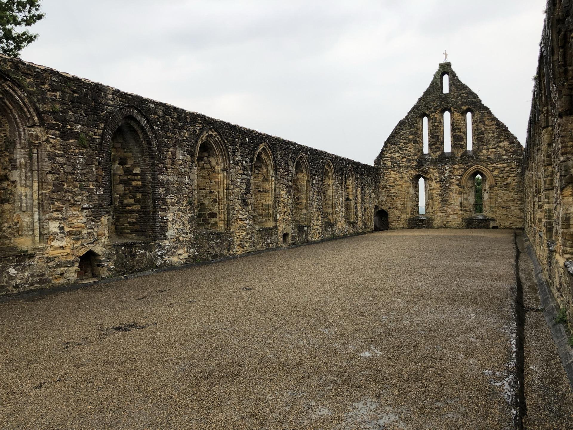 This image shows the ruins of a medieval abbey or church. The structure is characterized by a long nave marked by multiple arched windows along the walls, indicating a Gothic architectural style. At the far end, there is a more intact section with a pointed arch and several tall, narrow window openings that would have once held stained glass, leading to a roofless, arch-framed altar area. The ground is covered with gravel, and the remnants of the building give a historical and serene feel. The vegetation growing on and around the stone walls adds to the ancient and abandoned aesthetics of the place.
