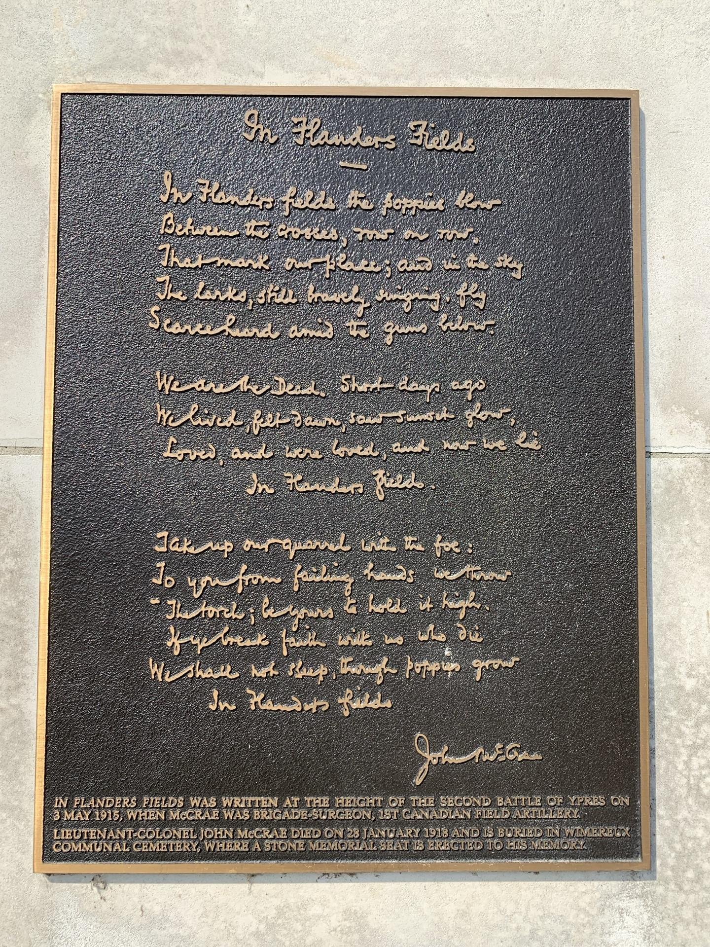 This image shows a plaque displaying the famous poem "In Flanders Fields" written by John McCrae. The plaque has embossed gold lettering on a dark background. Below the poem, there's additional text noting historical context:
- The poem was written during the height of the Second Battle of Ypres on 3rd May 1915.
- John McCrae was a brigade-surgeon of the 1st Canadian Field Artillery.
- McCrae died on 28th January 1918 and is buried in Wimereux Communal Cemetery, where a stone memorial seat is erected in his memory.

The poem itself articulates the experience of World War I and expresses mourning for the fallen soldiers, symbolized through the image of poppies growing between crosses marking the graves.
