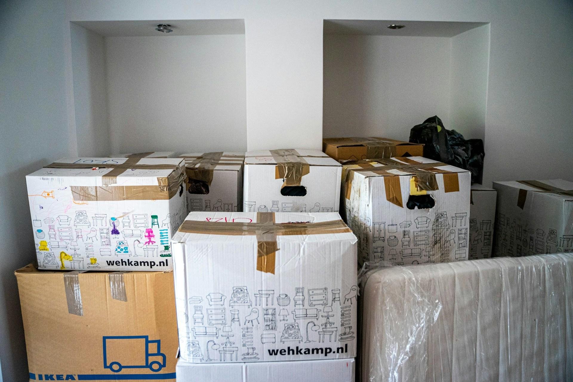 Cardboard boxes stacked in a room, some labeled with logos from IKEA and wehkamp.nl, with protective plastic covering furniture at the bottom right. The room appears to be in the process of packing or unpacking.