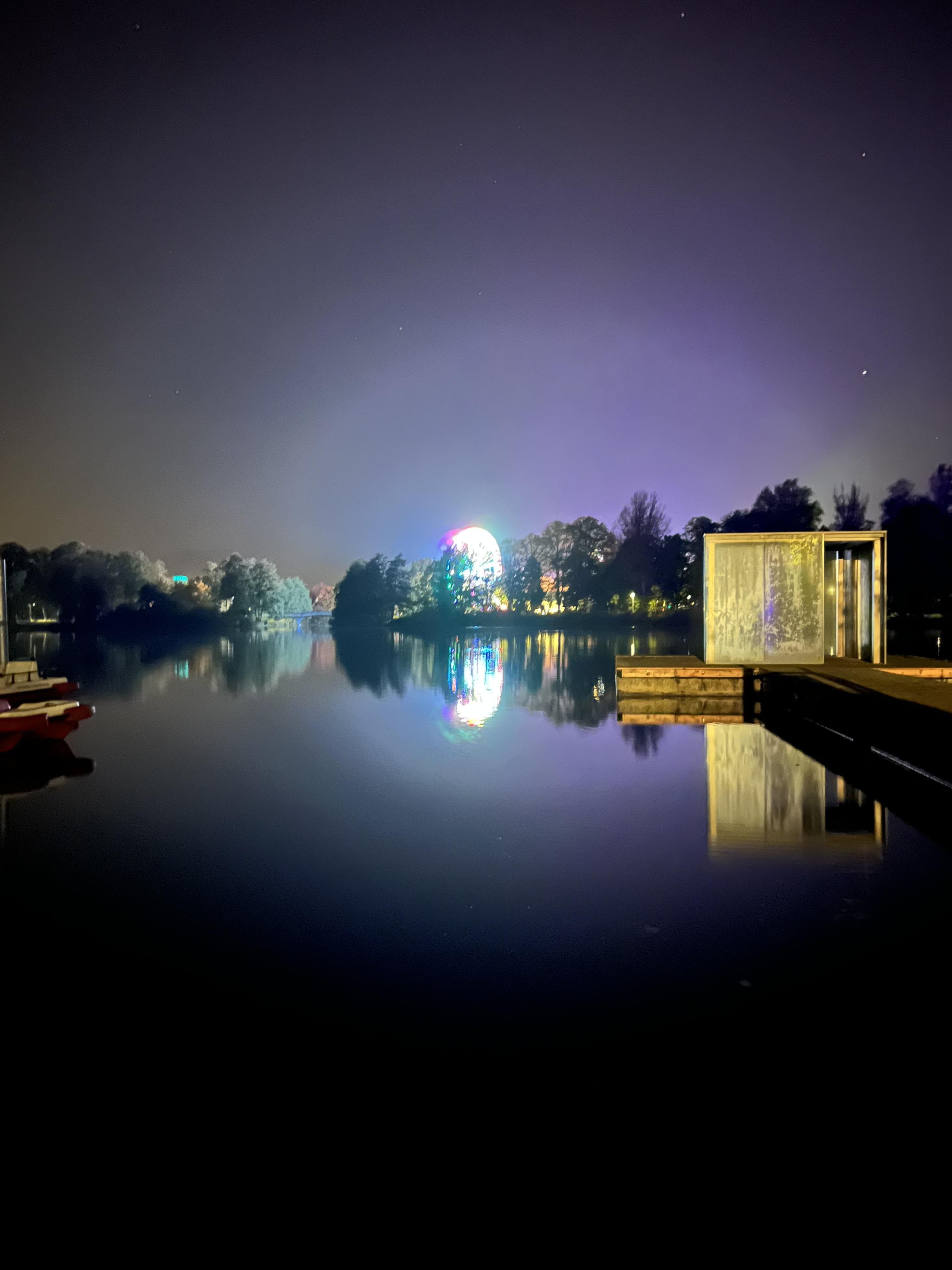 Nighttime scene over a still lake reflecting colorful lights. A luminous, multicolored Ferris wheel dominates the horizon amid silhouetted trees. Nearby, a dock with small boats and an illuminated glass structure borders the water. Stars dot the dark sky above.