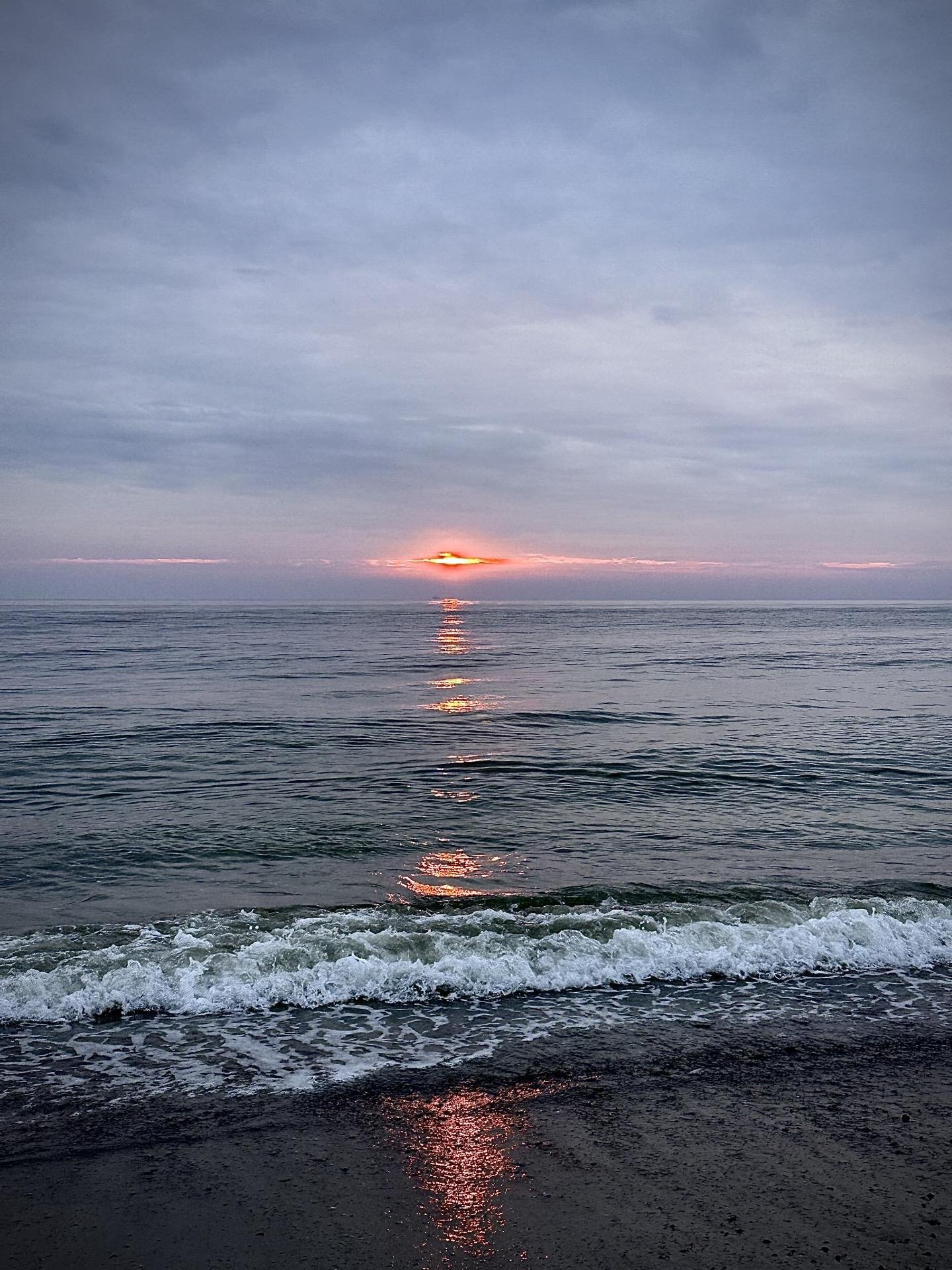 This image captures a serene beach scene at sunset. It features gentle waves coming onto the shore, with the sun partially visible as it sets on the horizon, casting a warm reflection on the water. The sky ranges in color from pale blue to shades of pink and orange near the disappearing sun, creating a peaceful and picturesque setting. The beach appears to be tranquil, without any visible people or significant activity, emphasizing the natural beauty of the seascape.