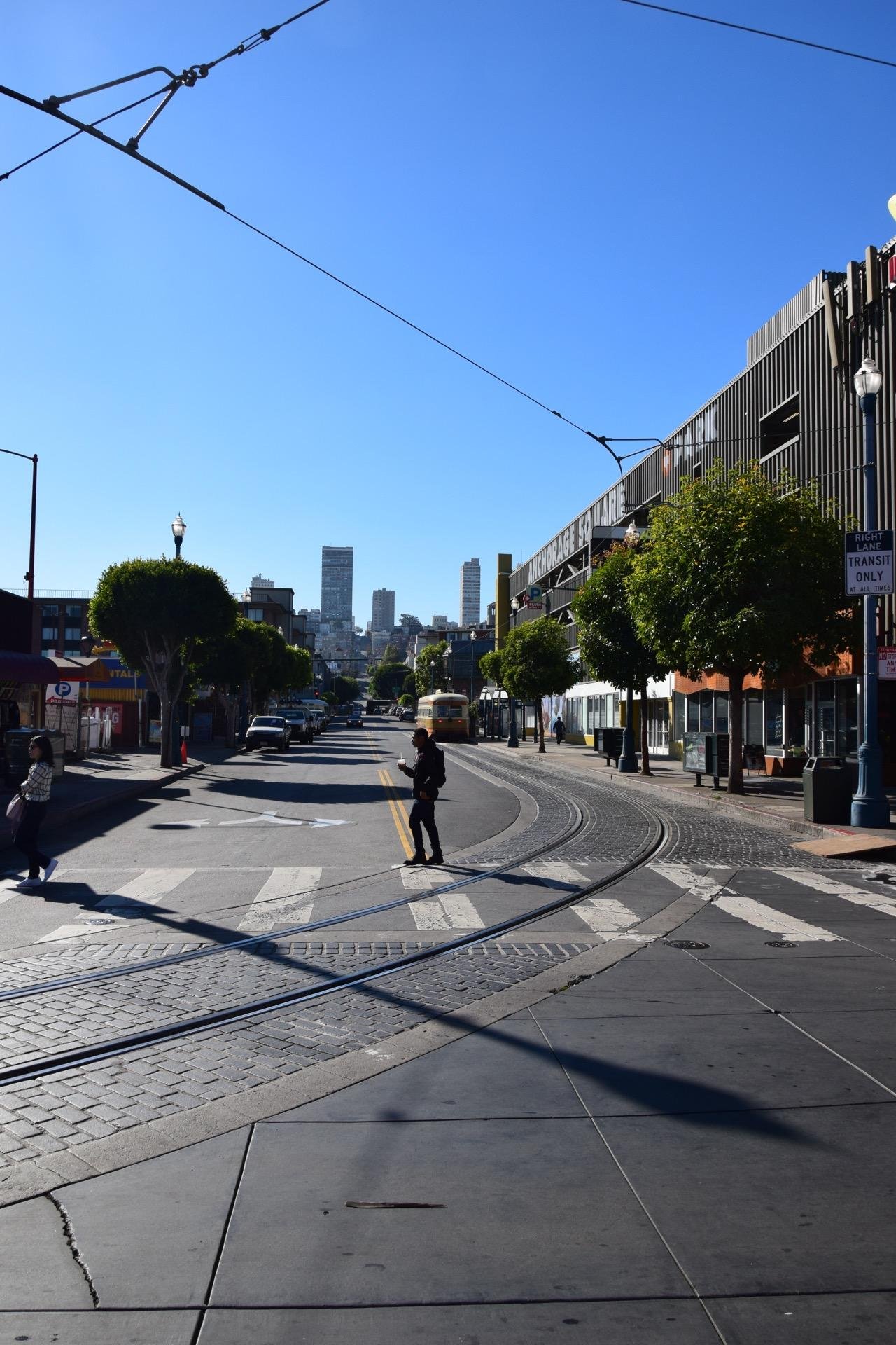 Street view depicting curved tram tracks and pedestrian crossing on a sunny day. A man crossing the street, people walking on the sidewalks, parked vehicles, and a tram in the distance are visible. The background shows a city skyline with high-rise buildings. Prominent signs and overhead wires are also featured. The scene conveys an urban environment, likely in a downtown area.