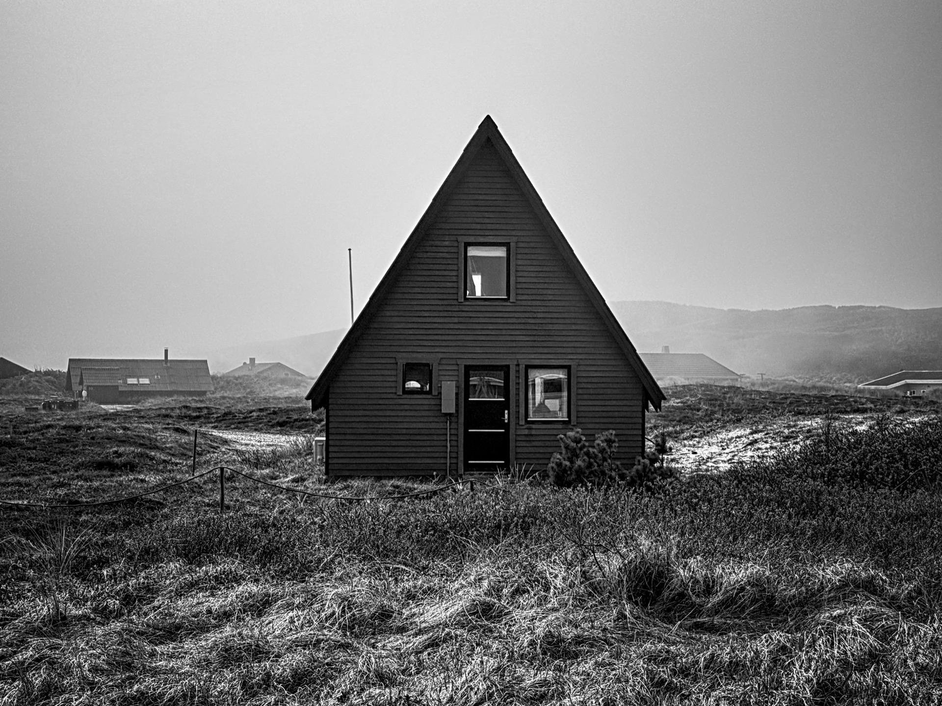 Black and white photograph of a triangular-shaped, dark-colored house centered in a grassy field with sparse vegetation. The house has a visible door and several windows, including a small one near the roof apex. Other buildings are partially visible in the foggy background, and a faint outline of mountains can be seen in the distance.