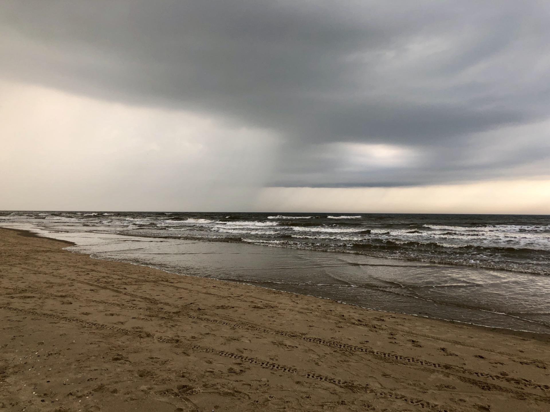 Overcast sky above a beach with choppy waves and dark clouds on the horizon; sandy beach foreground marked by tire tracks.