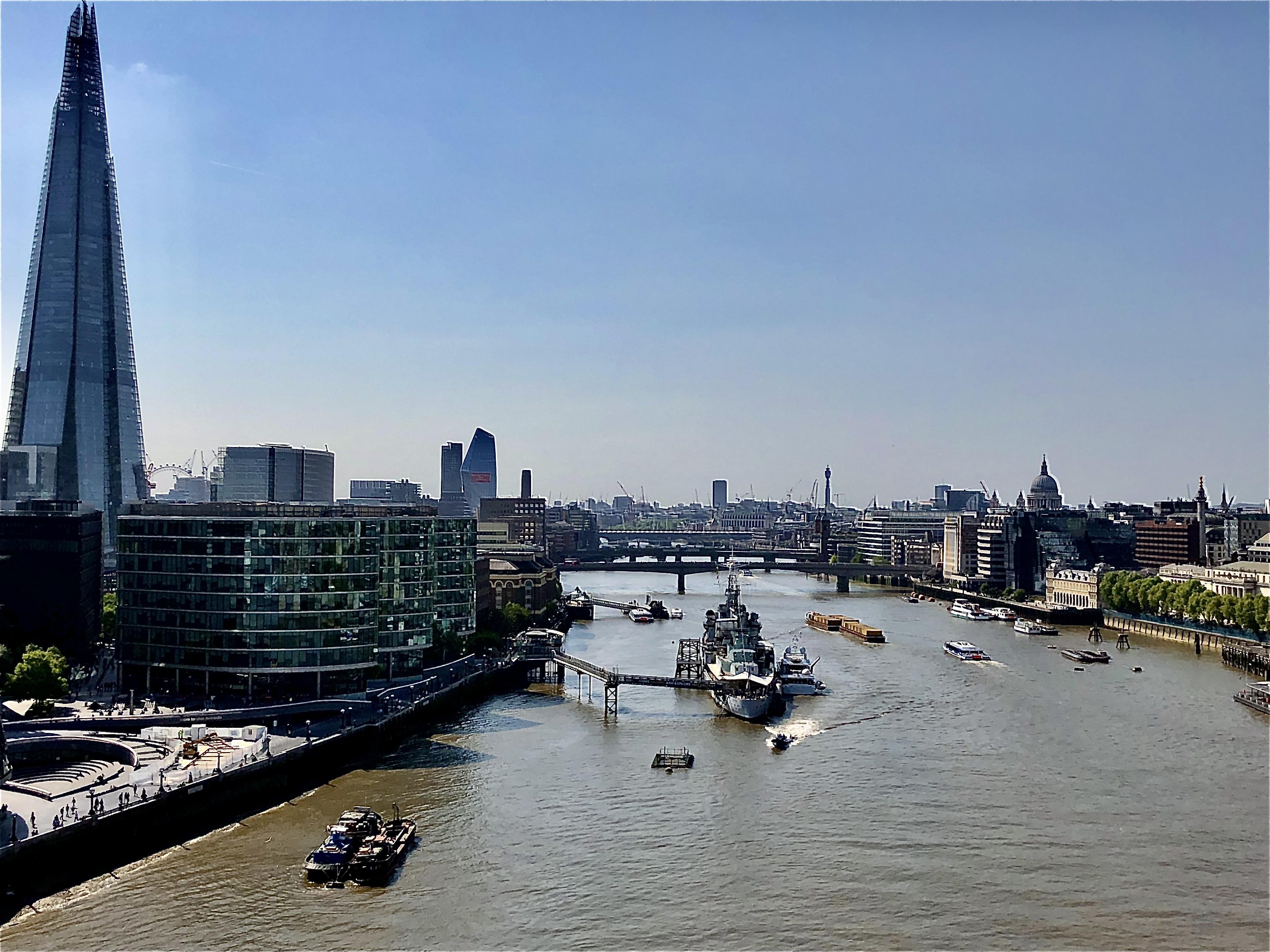 Aerial view of the River Thames in London, featuring busy water traffic including boats and barges. The Shard skyscraper dominates the left side, while various other modern buildings line the riverbanks. Multiple bridges span the river, and the cityscape includes recognizable silhouettes such as St. Paul's Cathedral in the distance under a clear blue sky.