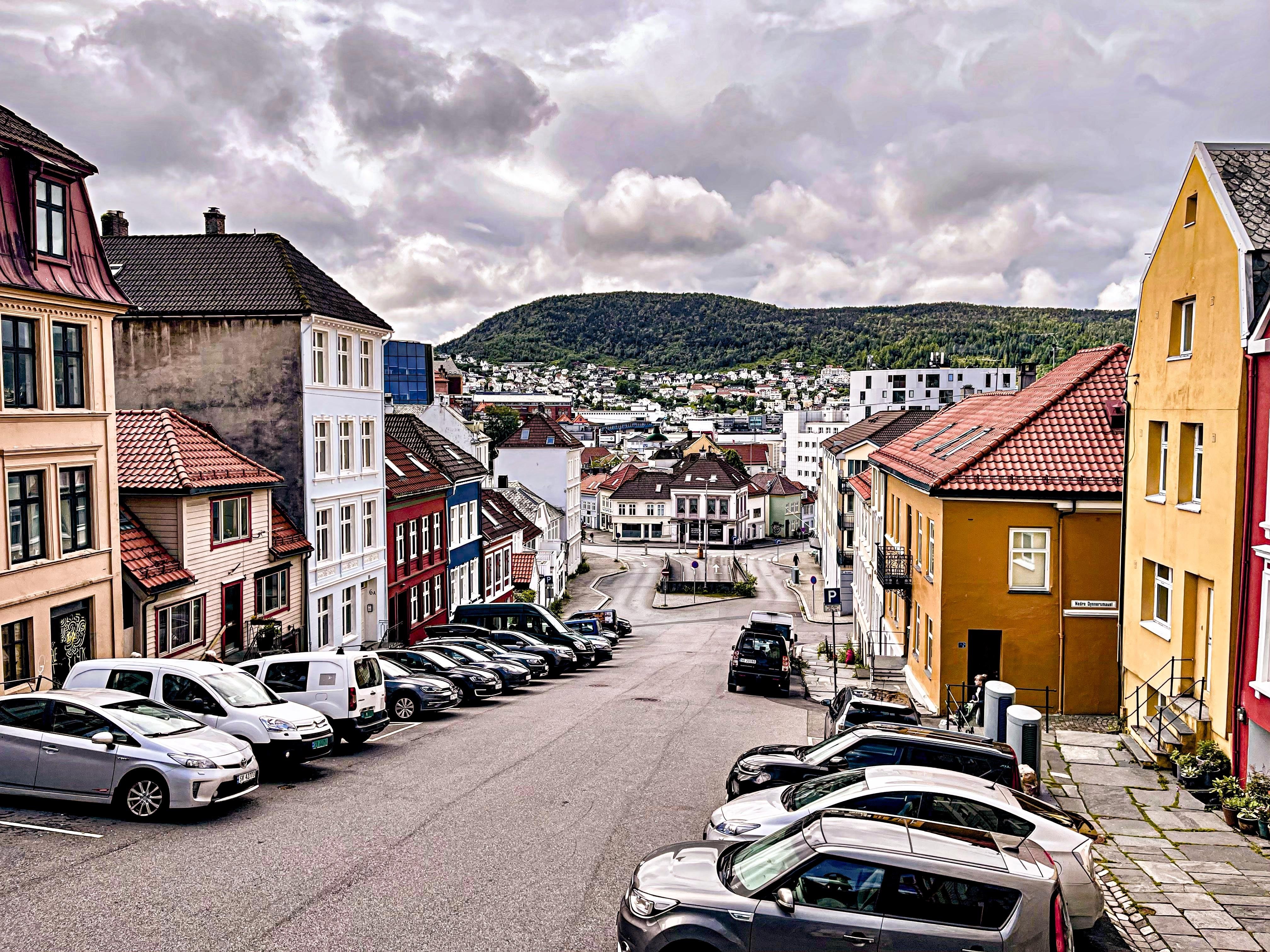 Steep street lined with colorful traditional houses leads down to a small open area with more buildings, overlooked by a forested hill in the distance. Cars parked along both sides of the street under a cloudy sky.