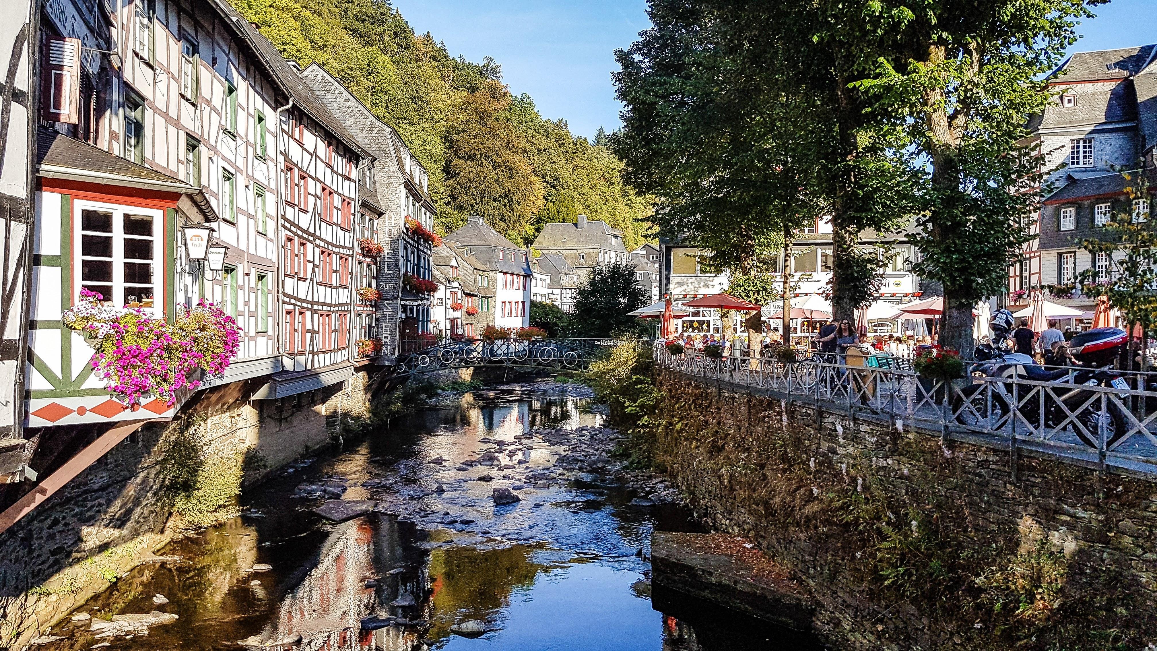 Half-timbered buildings with vibrant facades line a narrow river, reflected partially in the water. A pedestrian bridge crosses over the river, adorned with flower baskets. People are visible dining at outdoor tables under umbrellas near the riverbank. Trees and a clear sky complete the scene.