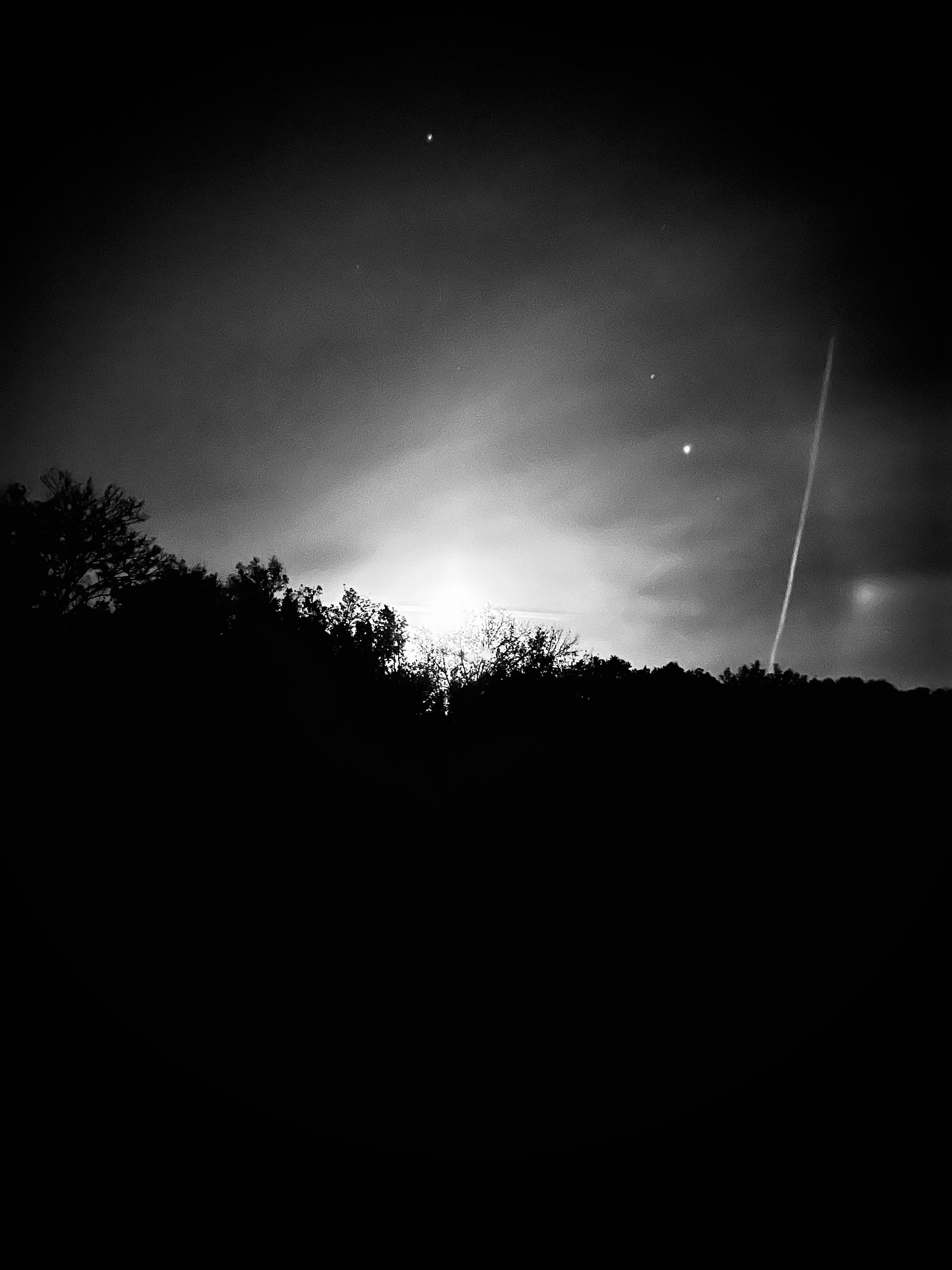 Black and white night sky with a bright moon partly obscured by a tree line, three visible stars, and a streak of an airplane trail on the right.
