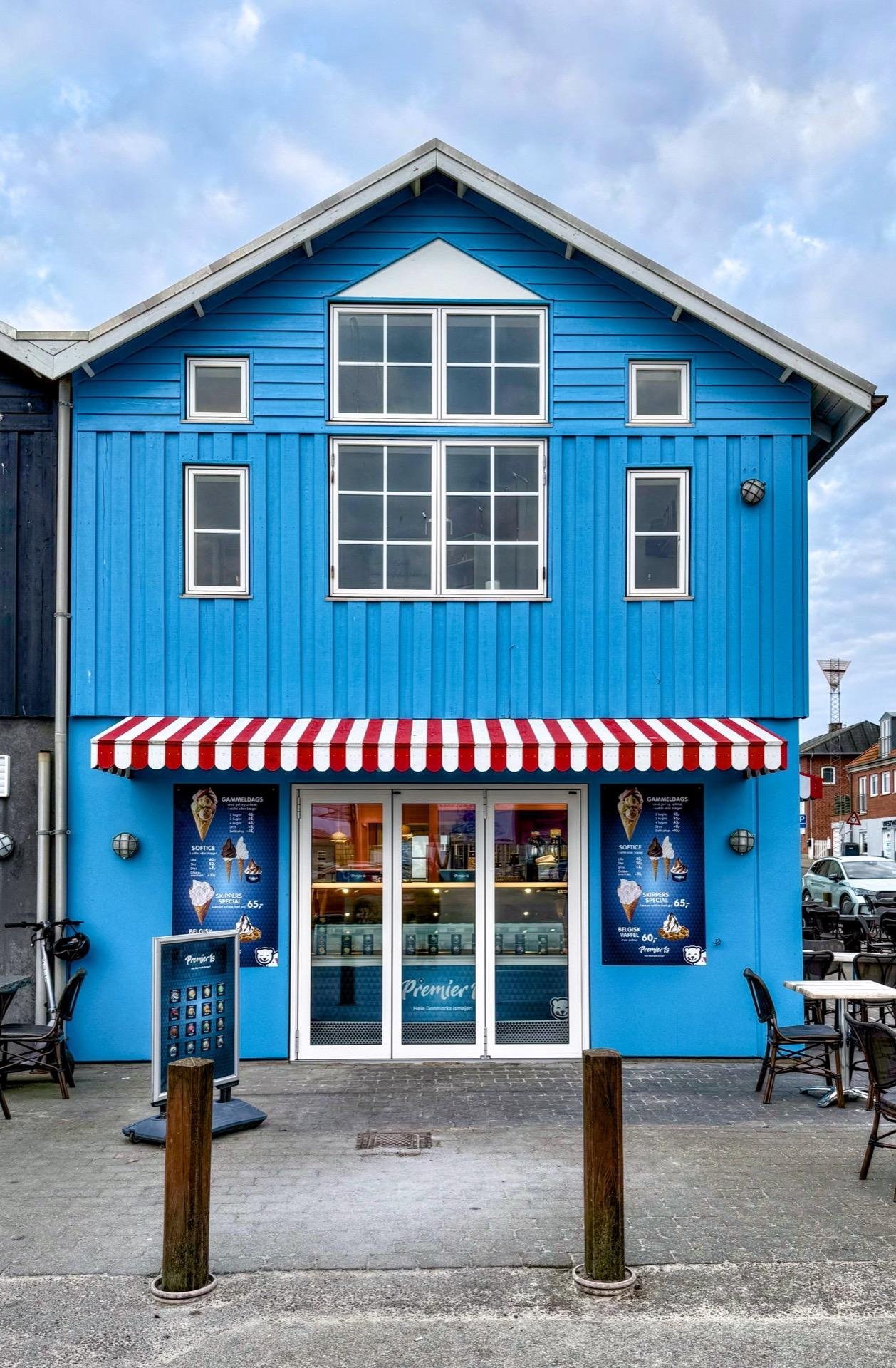 Bright blue two-story building with a gabled roof and multiple windows. The ground floor features a storefront with red and white striped awning and signage advertising ice cream, flanked by two wooden posts. Outdoor chairs and a table are visible to the side.