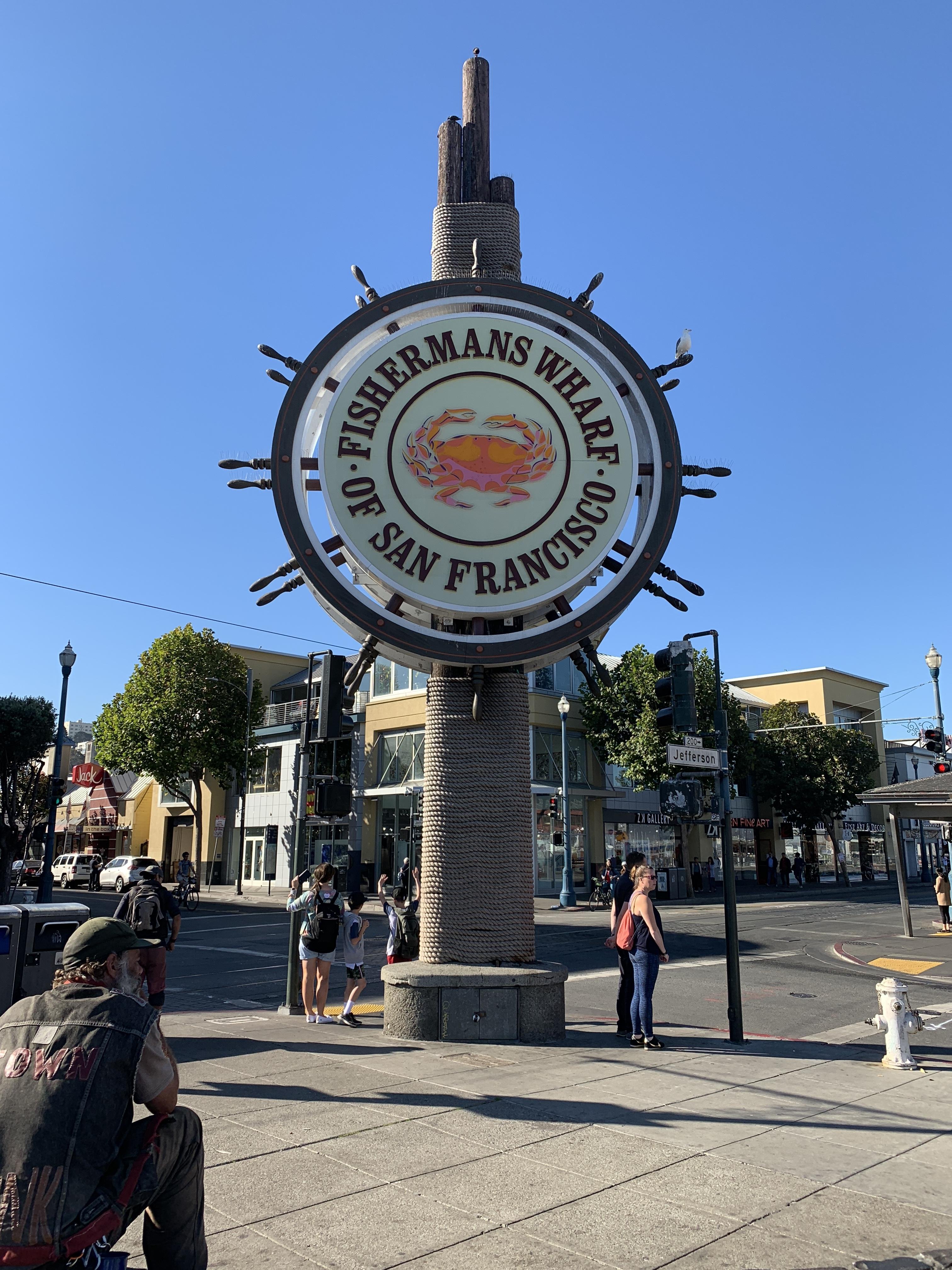 Circular sign reading "Fisherman's Wharf of San Francisco" mounted on top of a tall, textured pillar in an urban street setting. Below the sign, a person seated wearing a dark jacket and jeans captures the scene on camera. Other pedestrians are visible in the background near street corners and storefronts. Clear skies and sunlight illuminate the scene.