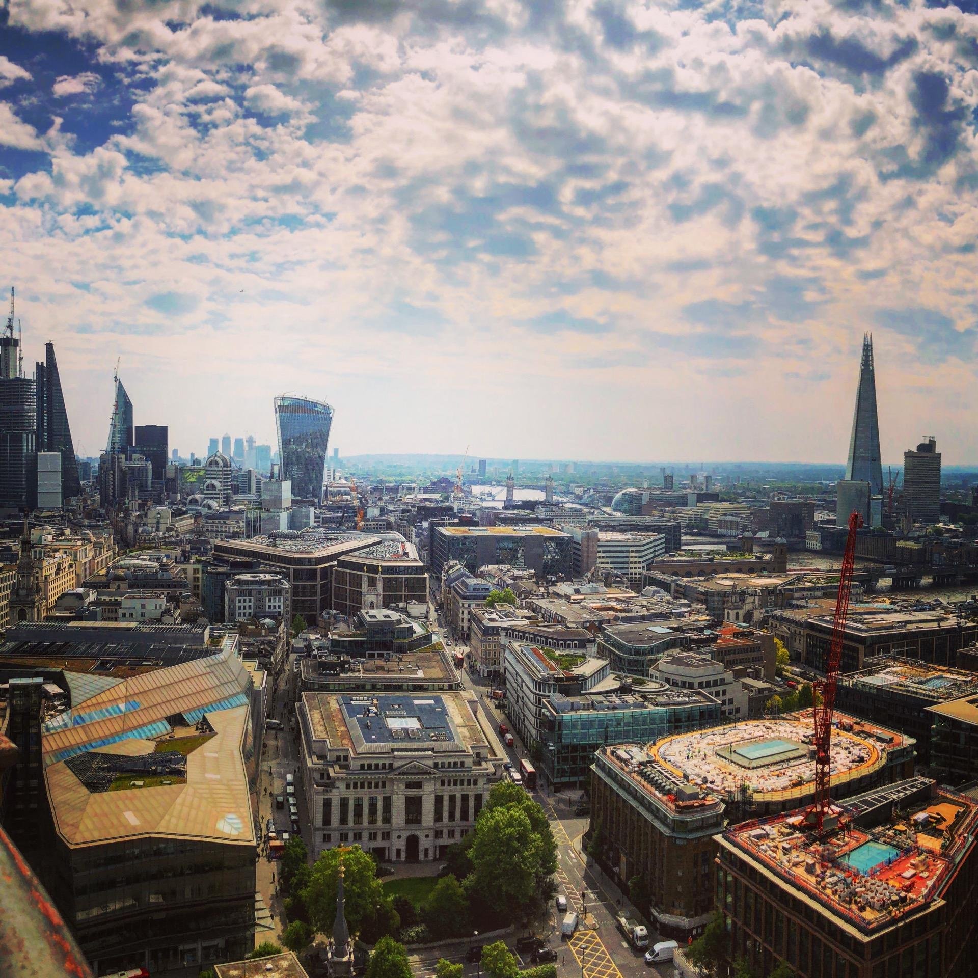 Aerial view of a dense urban cityscape under a partly cloudy sky, featuring a mix of modern high-rise buildings including distinctive skyscrapers, such as a cone-shaped tower and a wide curved rooftop, alongside traditional flat-roofed buildings. The foreground shows active construction sites, city streets, and vehicles, with a lush green backdrop in the distance extending to the horizon.