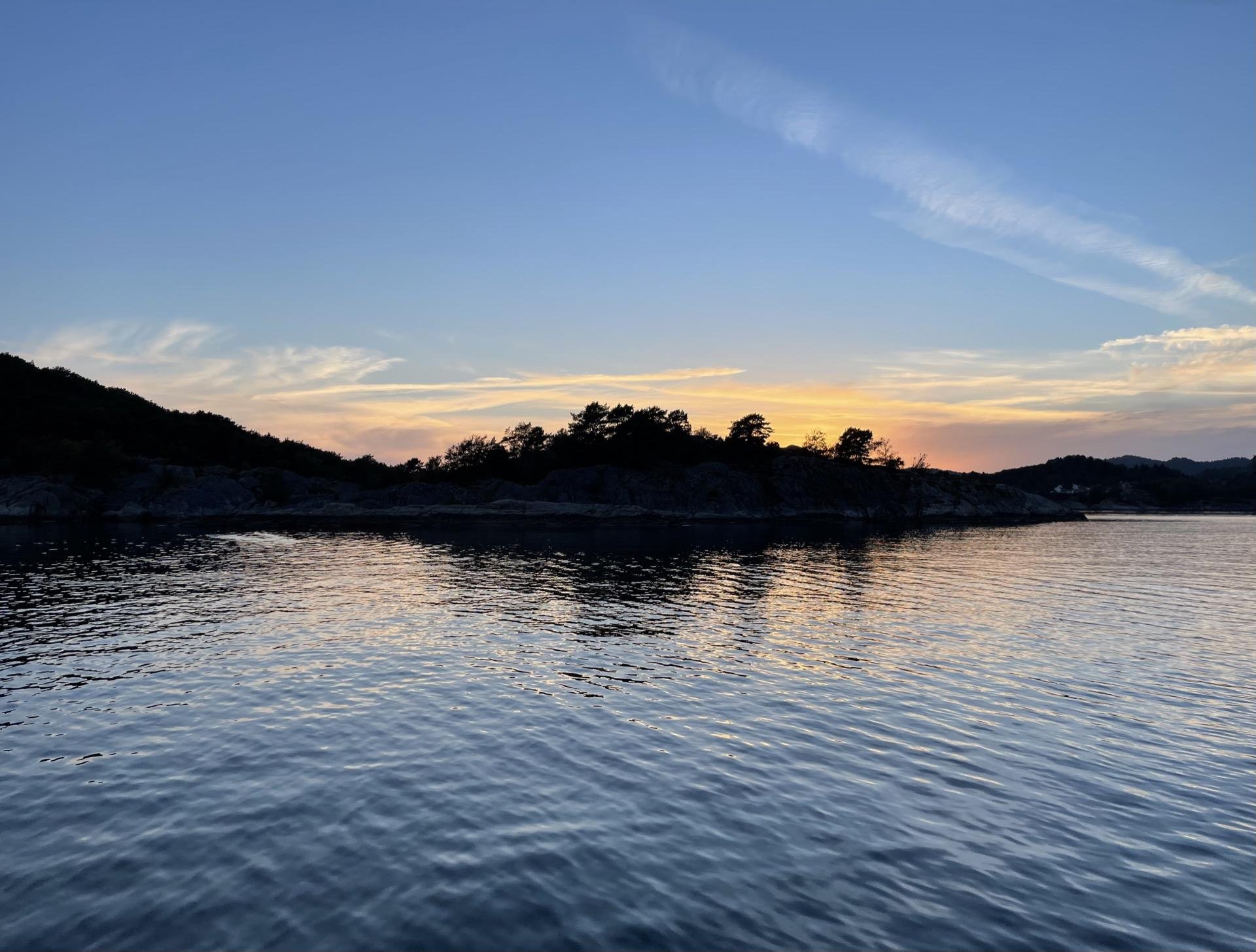 Sunset over a calm sea with the silhouettes of rocky shores under a clear sky with gradations of blue to orange near the horizon. Light reflections shimmer on the water's surface.