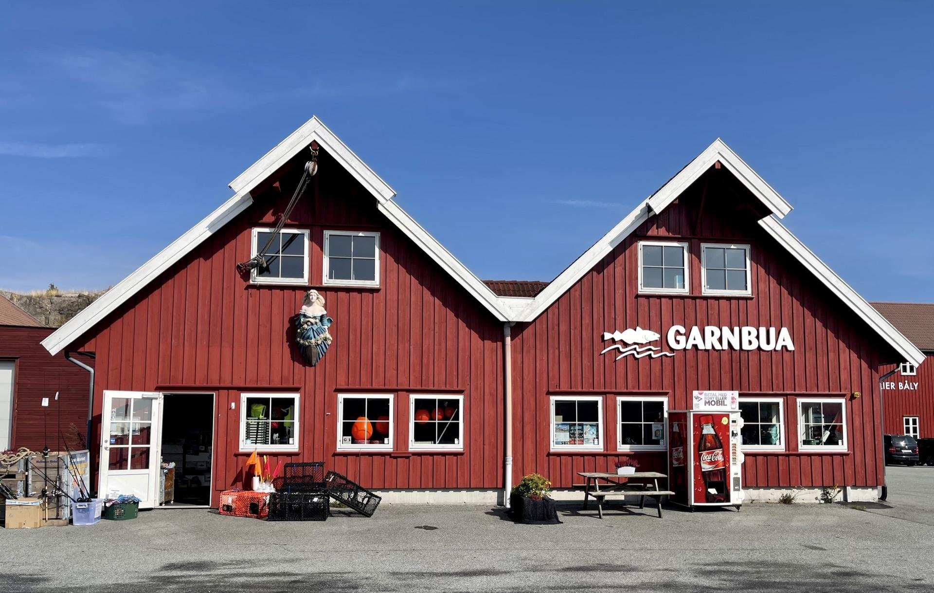Red wooden building with symmetric peaked roofs and central ornament of a mermaid. Labeled "GARNBUA," the facade features two large windows and double doors, flanked by objects including a soda vending machine and assorted maritime decor. A clear blue sky forms the background.