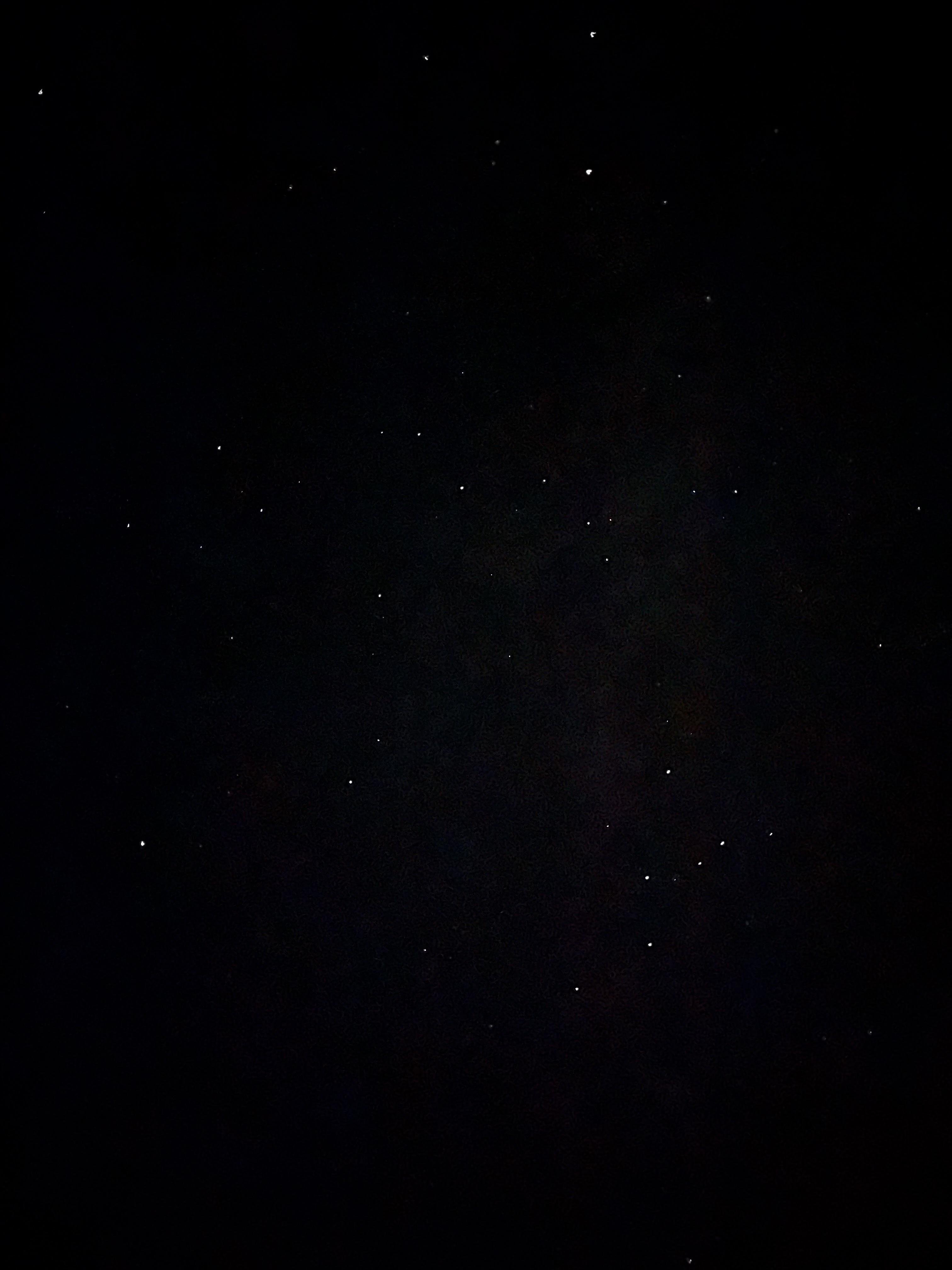 A dark night sky scattered with numerous small, bright stars. The majority of the stars appear white, with a few exhibiting a slight yellow or blue hue. The background is mostly black, indicating low light conditions typical of nighttime sky observations.