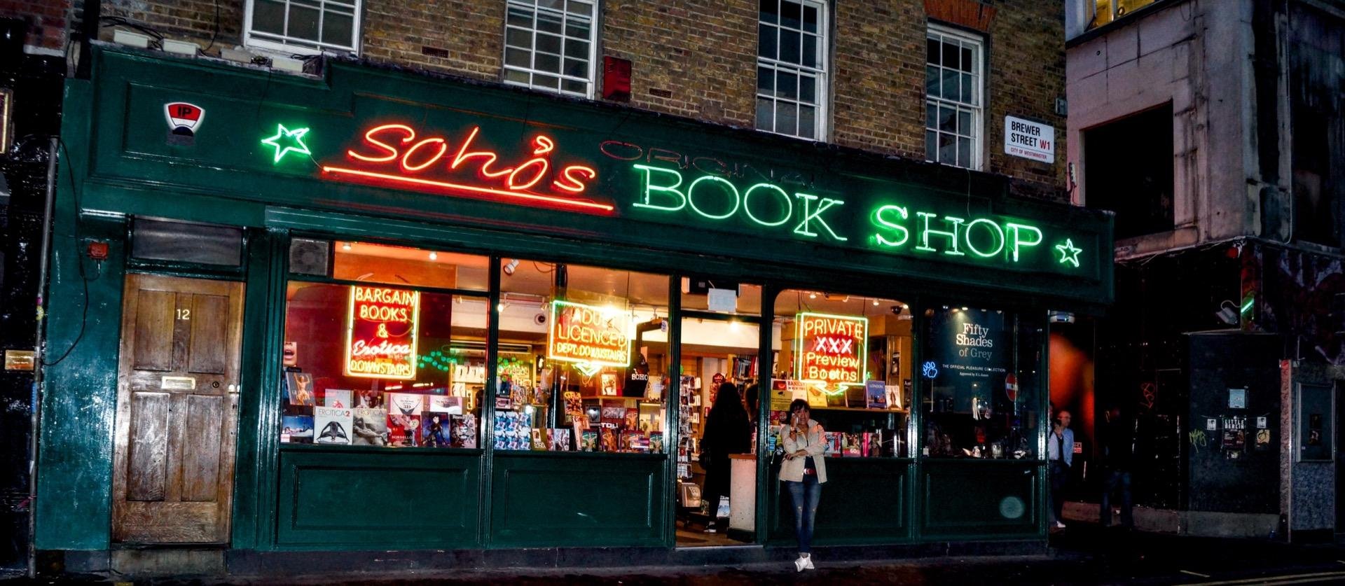 Nighttime view of "Soho's Book Shop" facade lit by neon signs, featuring a green storefront and large, brightly lit display windows filled with books and posters. A person stands outside, slightly left of center, looking at the shop's window.