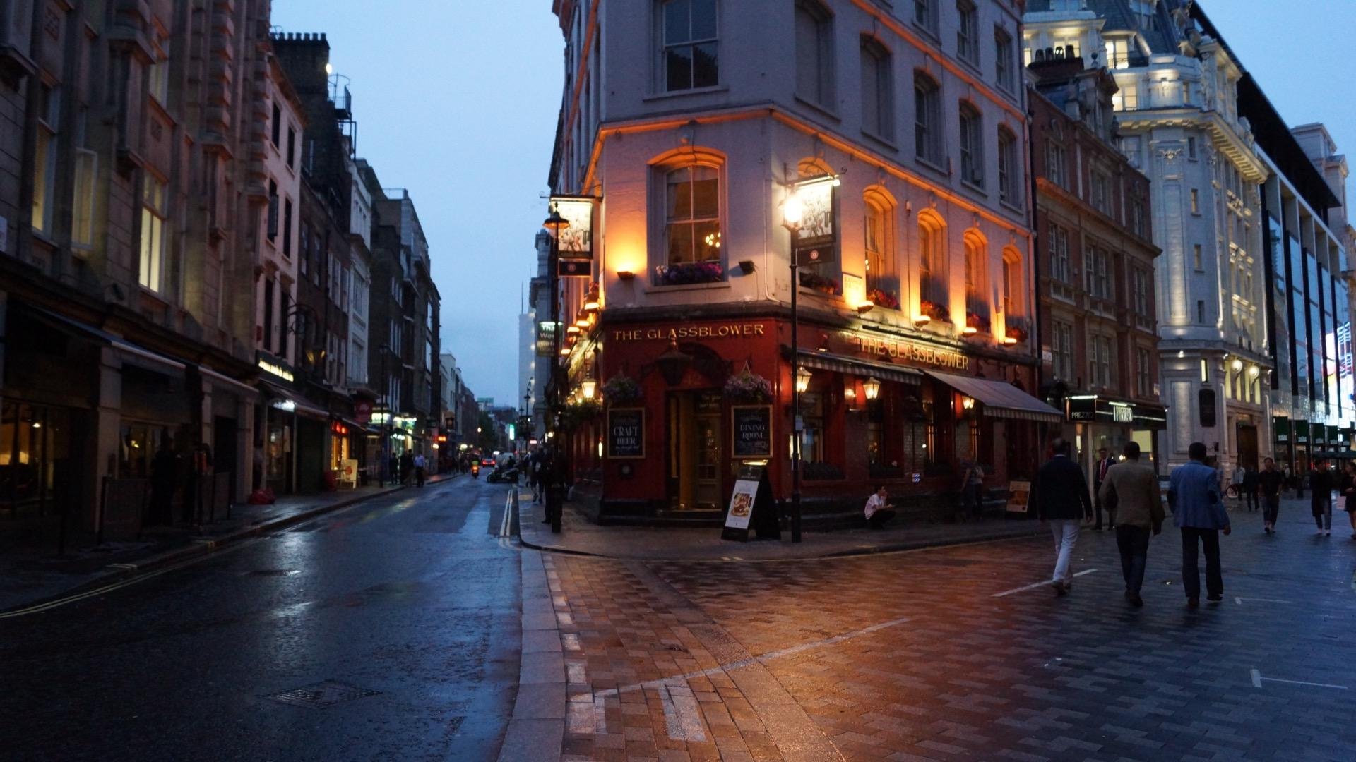 Twilight over a wet city street corner with a prominent red building labeled "The Glassblower" lit with warm lights. Sidewalks glisten under streetlamps, reflecting pedestrians and the dusky blue sky. Other buildings fade into the background.