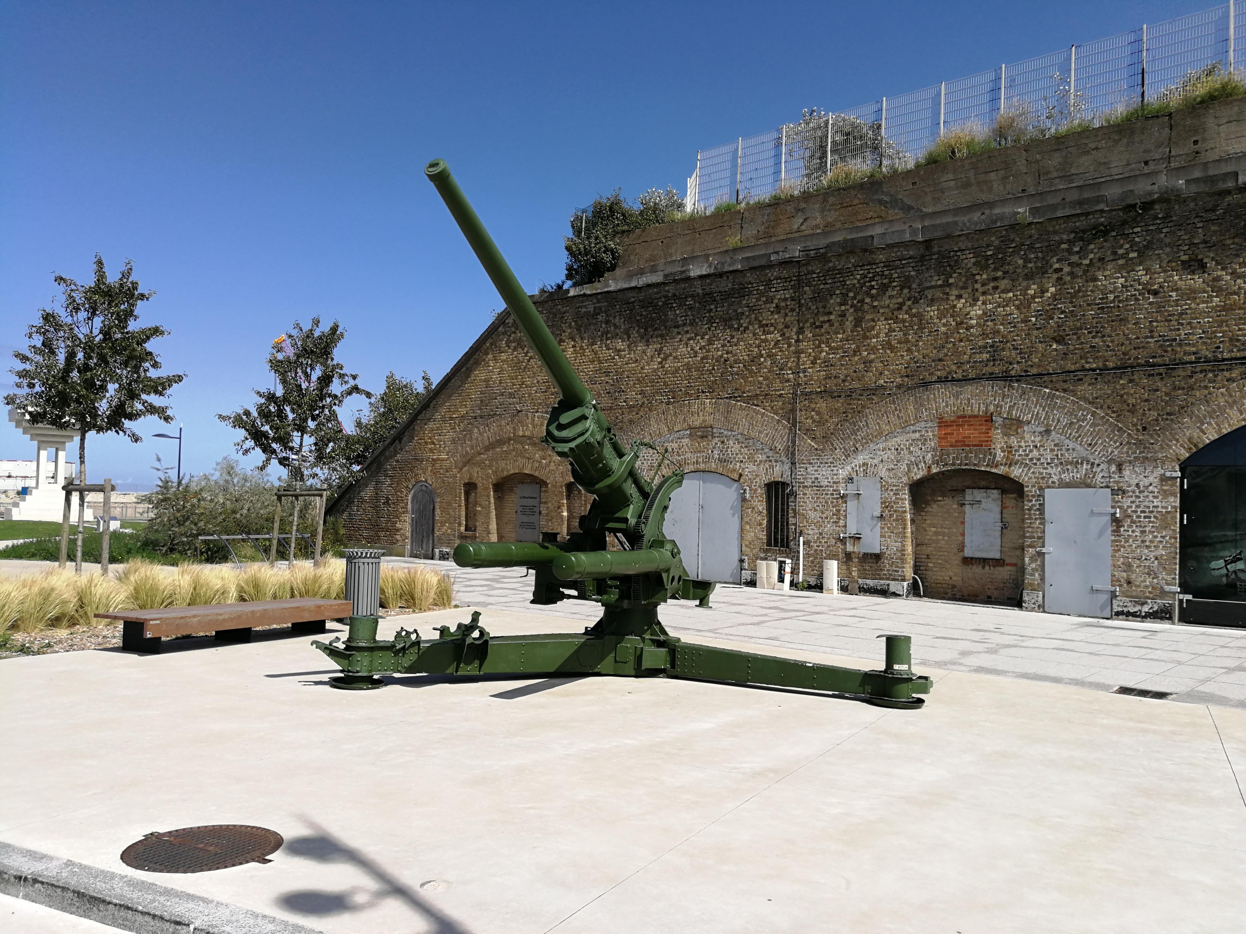 Large green cannon mounted on a rotating base, positioned on a grey concrete platform outdoors. Behind the cannon is an old brick building with arched doorways blocked by white doors and graffiti. Two trees flank the cannon, with a clear blue sky above and patches of grass nearby. A stone wall topped with a metal fence rises behind the building.