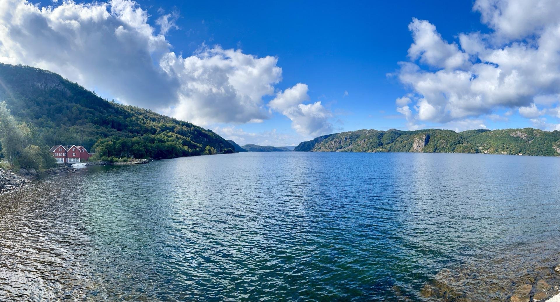 Panoramic view of a large lake bordered by mountains under a partly cloudy sky. On the left, a group of red-roofed houses nestles on the shore.