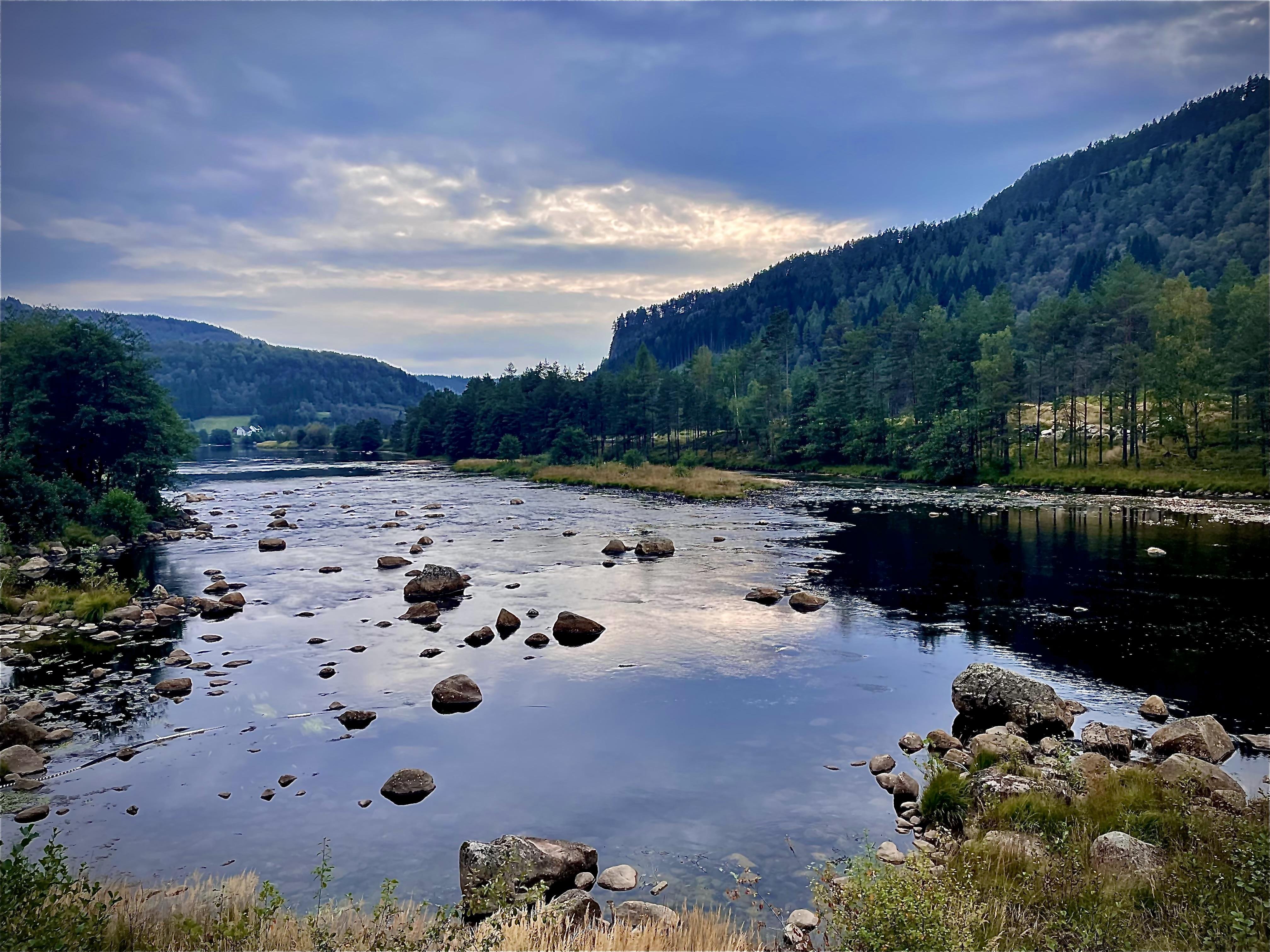A serene river landscape at twilight with scattered boulders in the gently flowing water. Dense forests border the riverbanks, highlighted against a cloudy sky. Distant hills contribute to a layered vista in shades of green and blue.