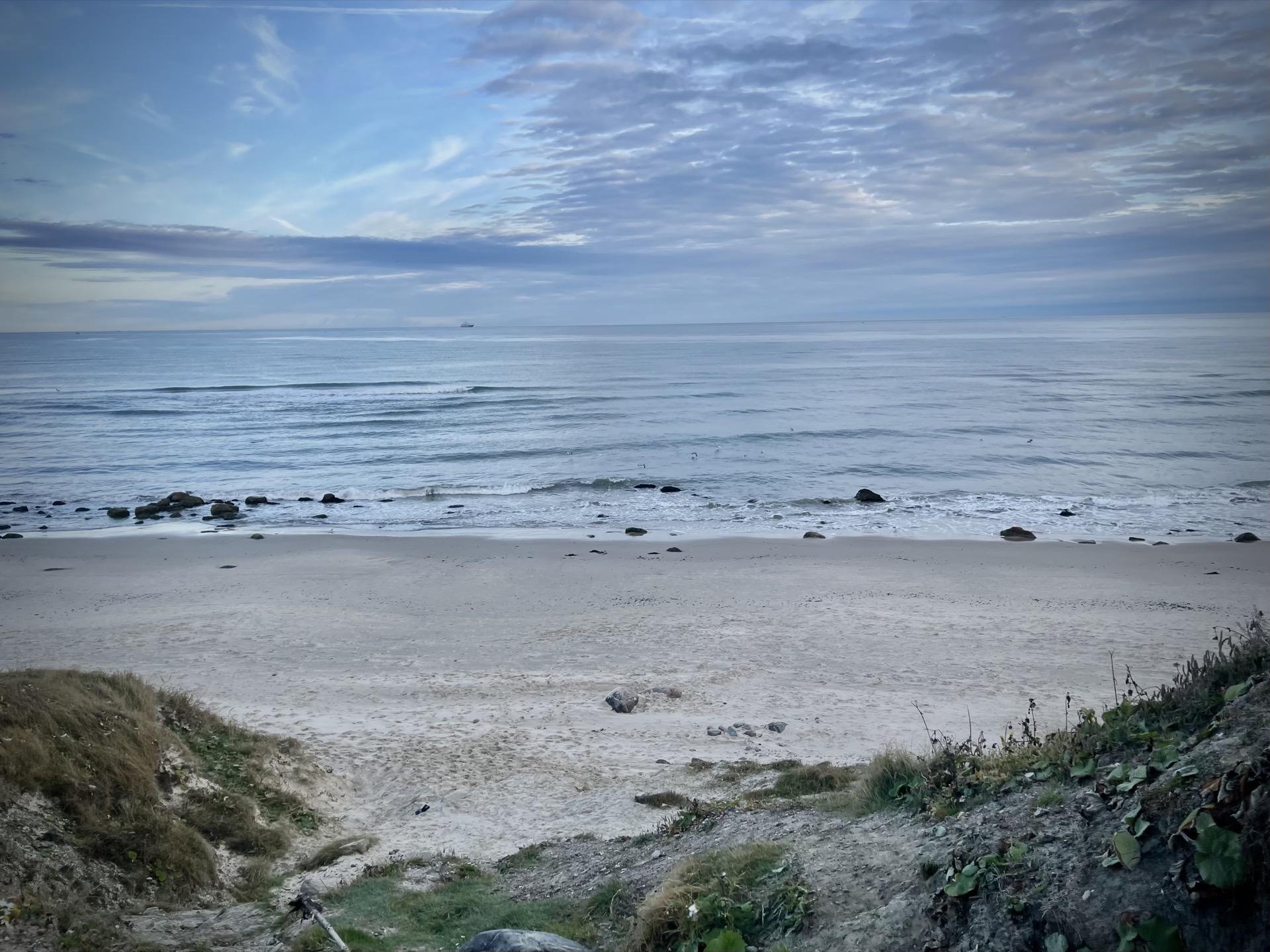 Sandy beach with scattered rocks leads down to a calm ocean under a cloudy sky at twilight. A grassy slope with a pathway appears in the foreground.
