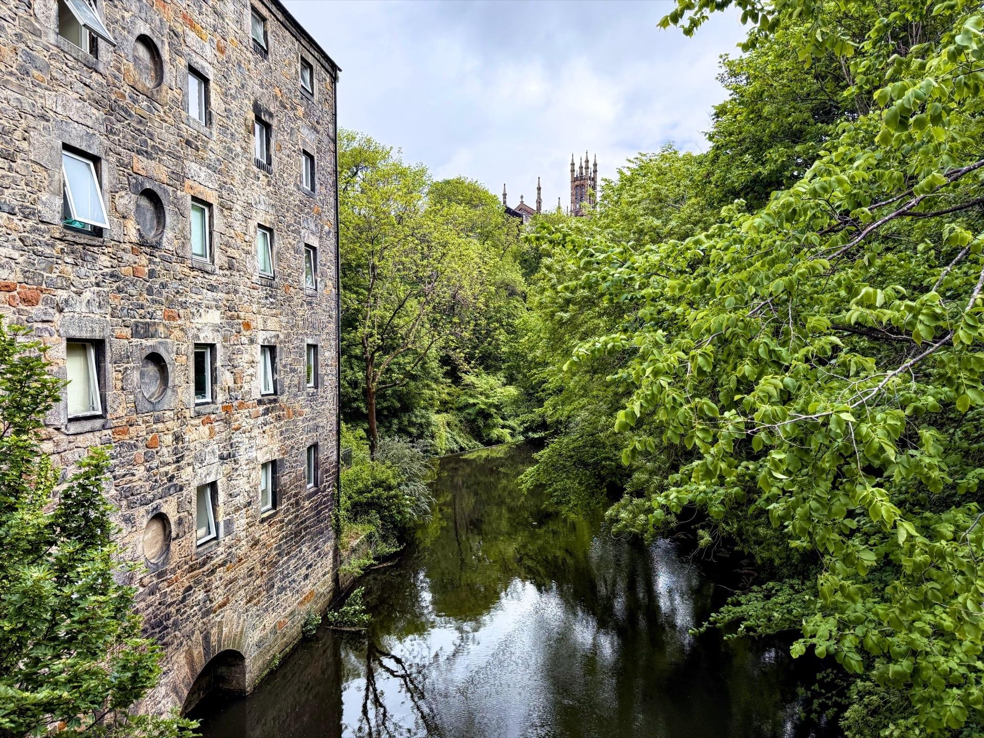 Stone building with arched ground-level openings beside a calm river, surrounded by dense green foliage. Gothic-style towers peek above the tree line in the background.