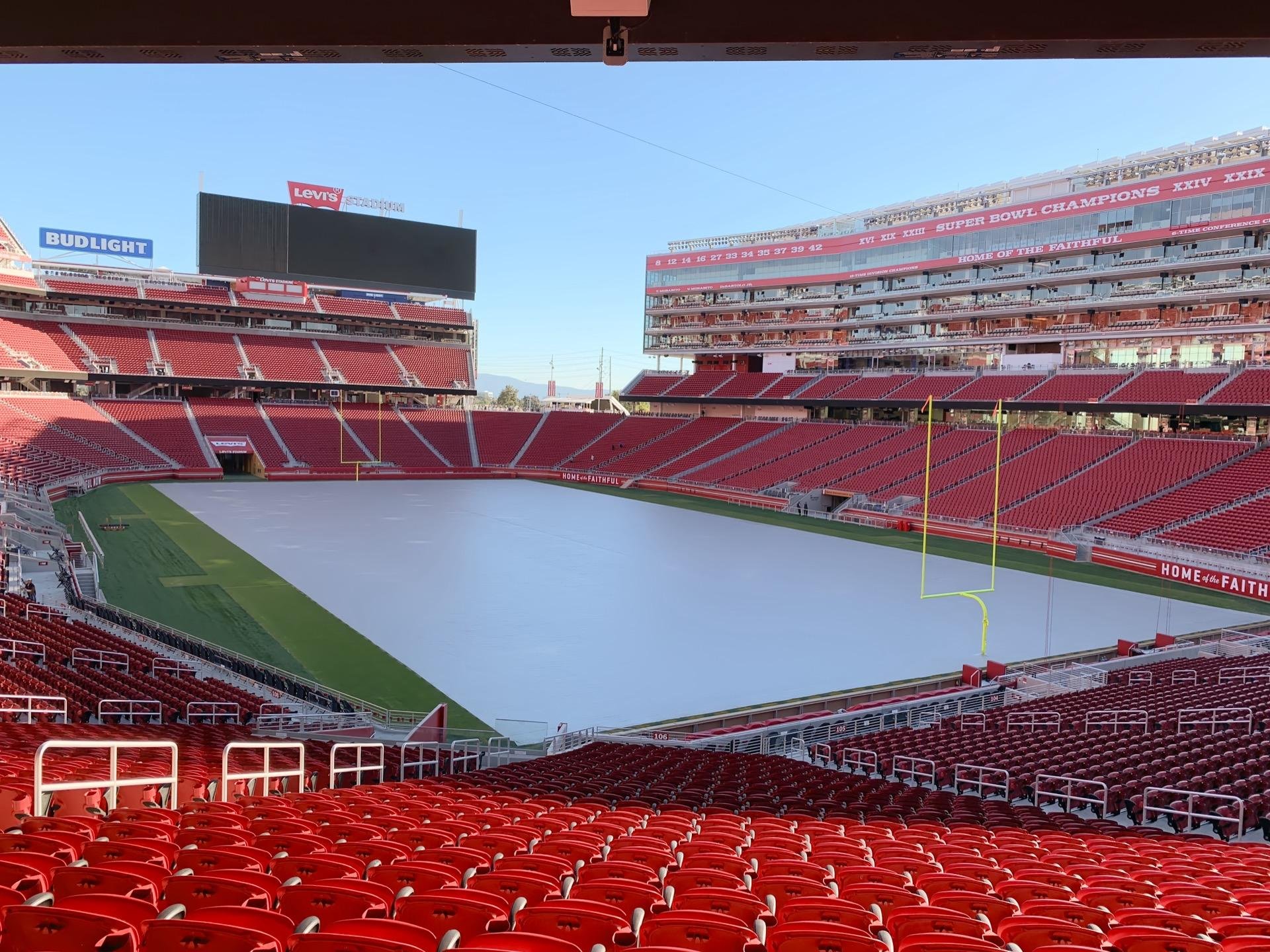 Empty football stadium with rows of red seats in the foreground and a large digital scoreboard at the far end. The playing field, bordered by green turf, is covered with a white tarp. Notable banners, including a Bud Light advertisement and a Levi's sign, dominate the top-tier stands, which also display text citing Super Bowl championships.