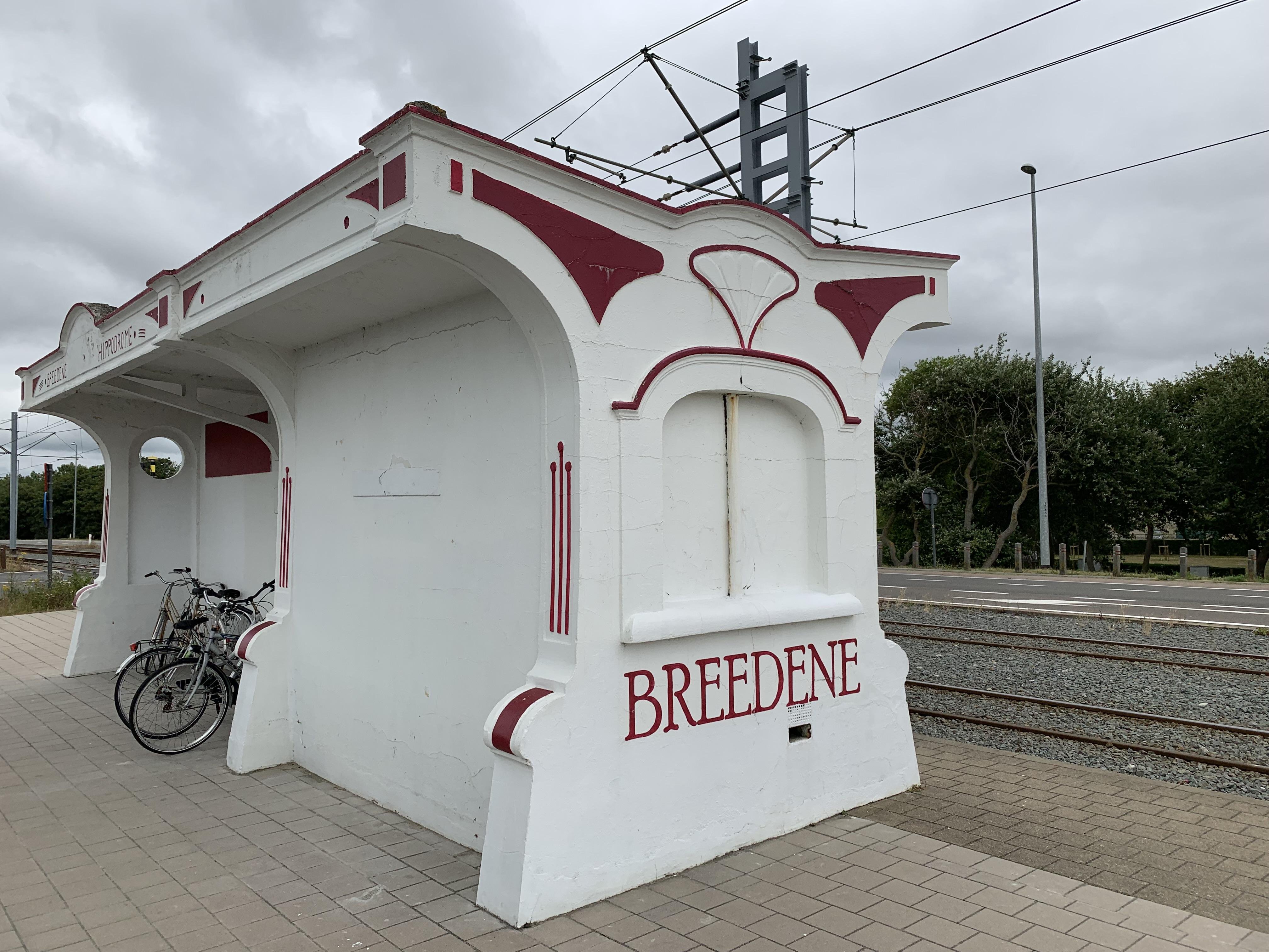 White and red train station shelter labeled "BREDENE" with arched openings and ornate decorations, situated beside railroad tracks. A bicycle is parked beside the shelter. Overcast sky and greenery in the background.