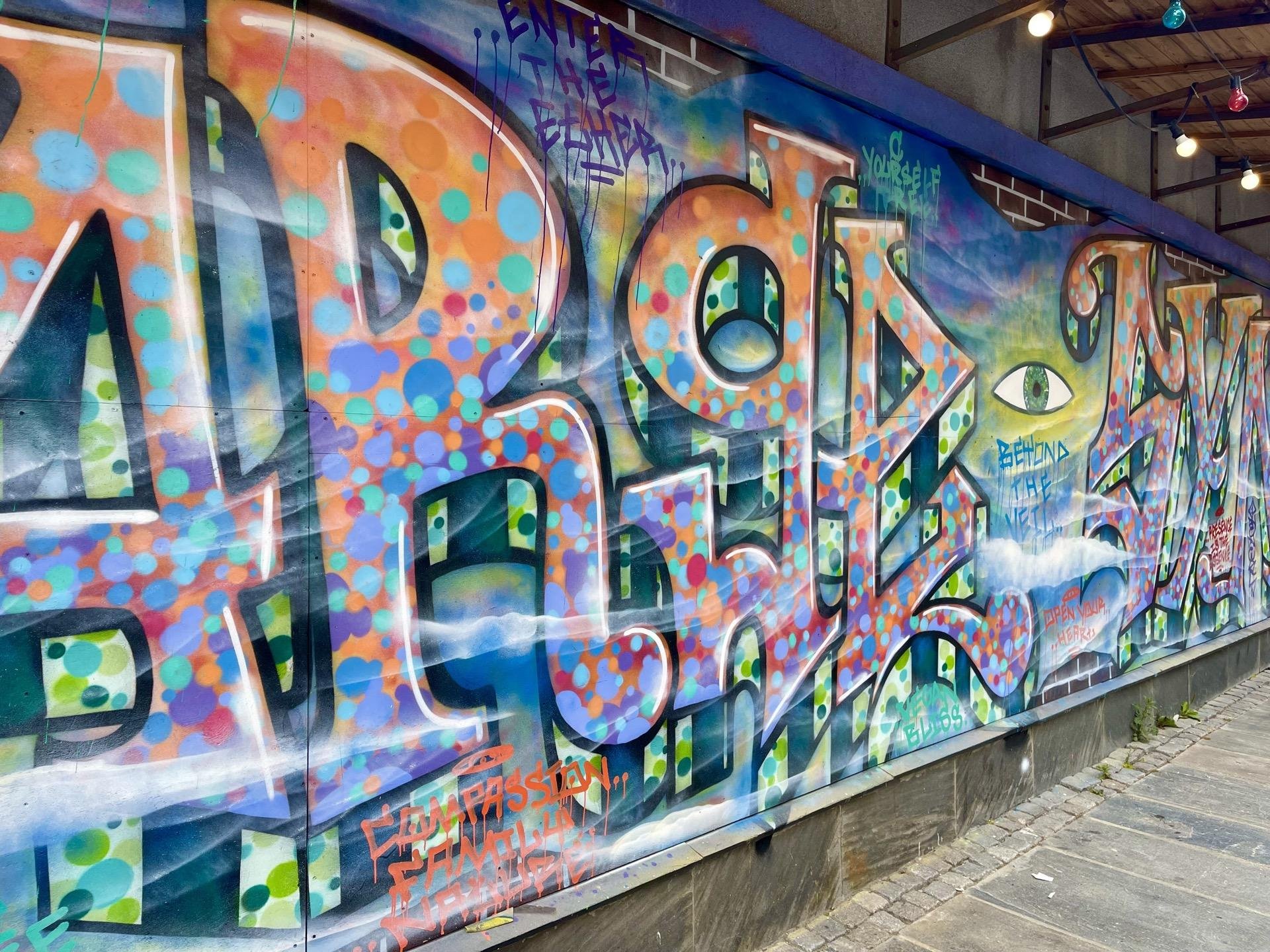 Colorful graffiti art covers a wall, featuring large, stylized letters in an array of colors with dot and splatter patterns, and a detailed eye incorporated into the design. The phrase 'compassion, family, yourself' is visible among the vibrant artwork.
