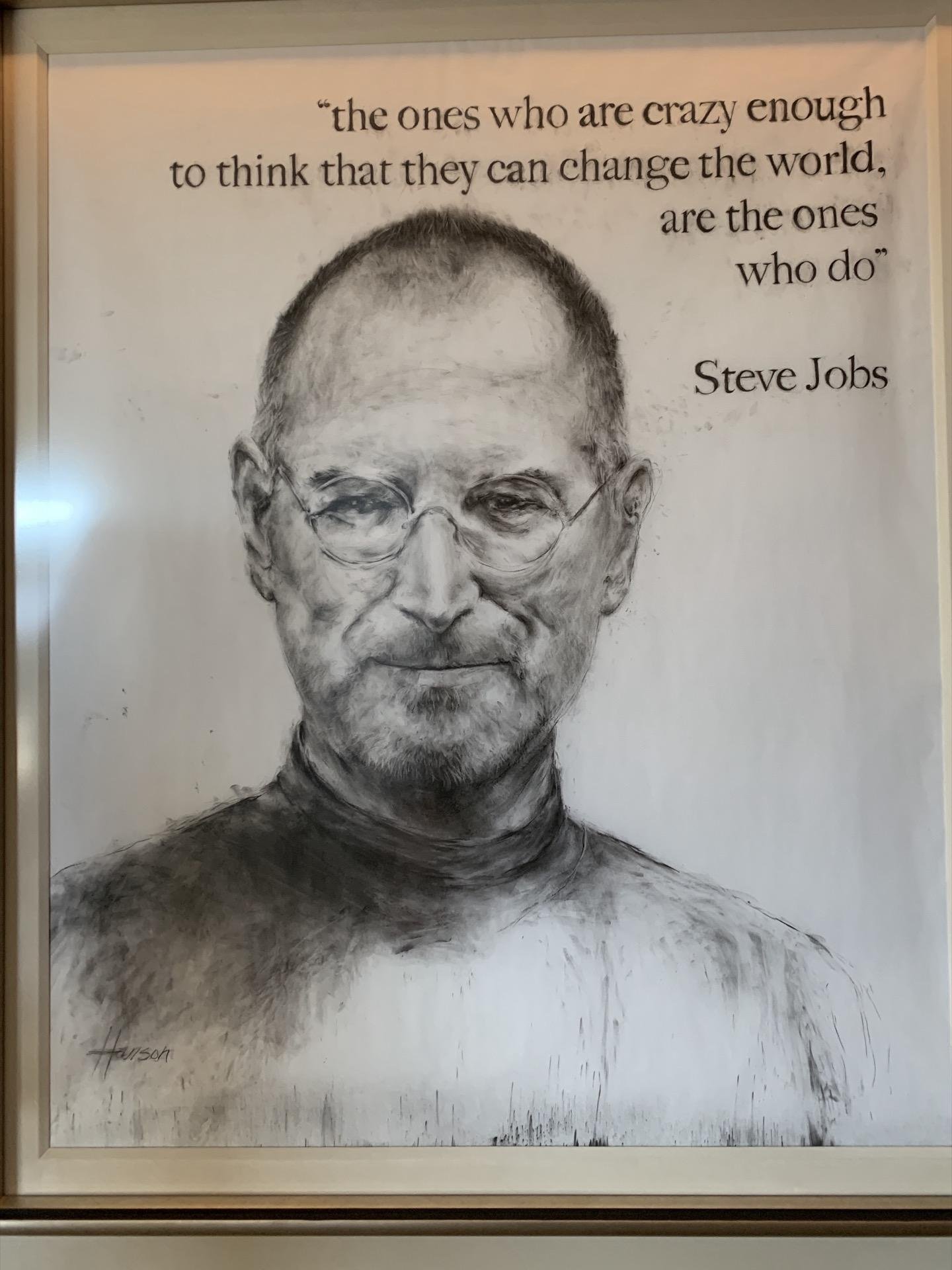 Black and white charcoal sketch of a middle-aged man wearing glasses and a turtleneck, framed and displayed on a white wall. Above the portrait is a quote attributed to Steve Jobs: “the ones who are crazy enough to think that they can change the world, are the ones who do.” The artist's signature is in the bottom left corner of the sketch.