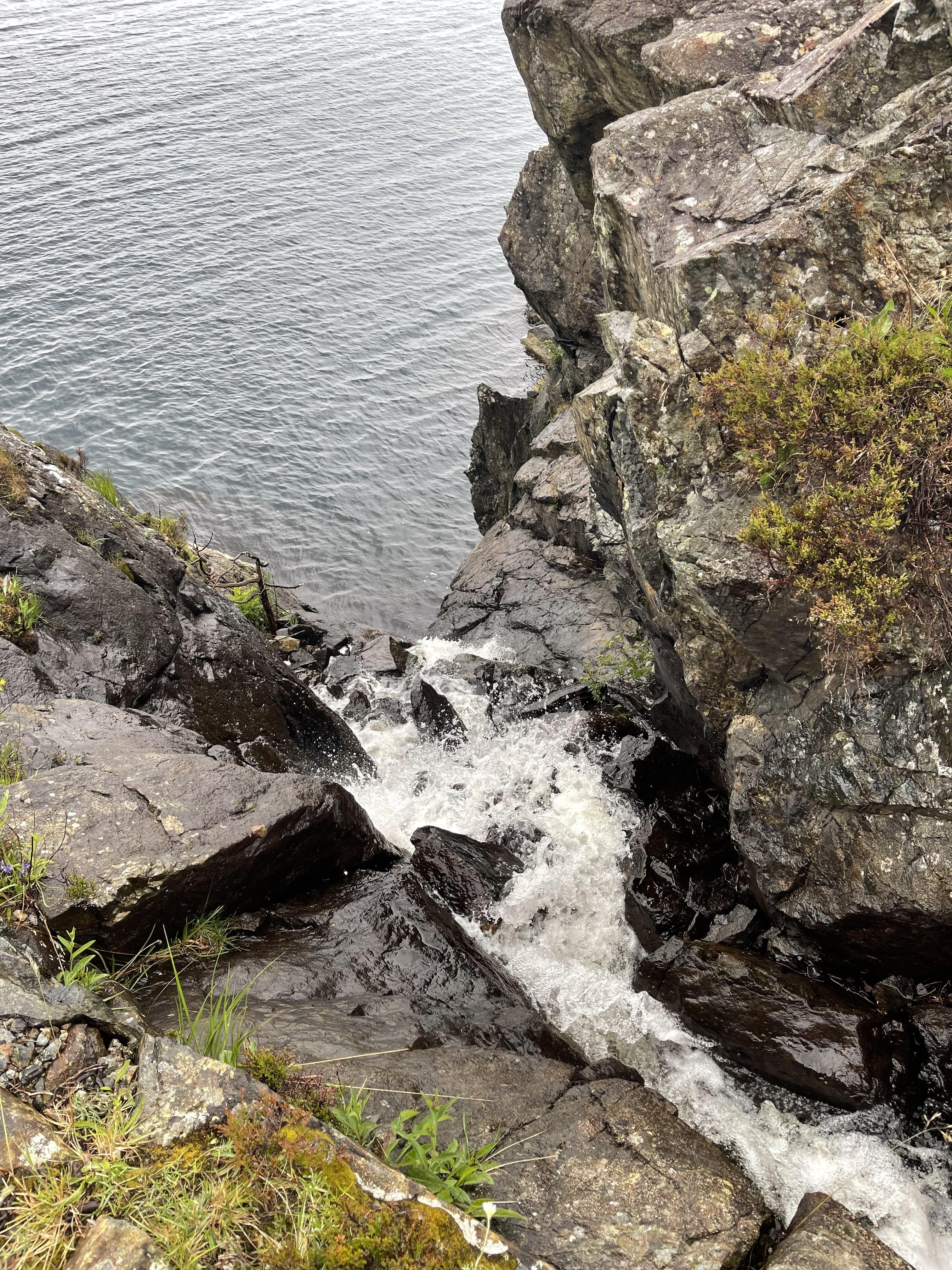 Vertical view of a rough, rocky cliff edge with a small waterfall cascading down into a calm, expansive body of water. The rocks, covered with patches of green moss and some grass, are wet and dark, suggesting recent water flow. The water appears frothy where it hits the base of the cliff before merging with the larger body of grayish water.