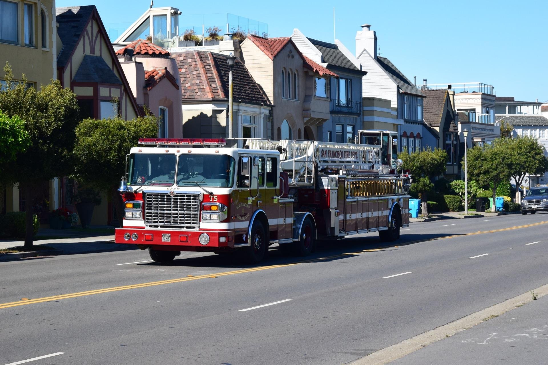 A red and white fire engine marked "San Francisco Fire Dept" travels on a city street with traditional houses on both sides under a clear blue sky.