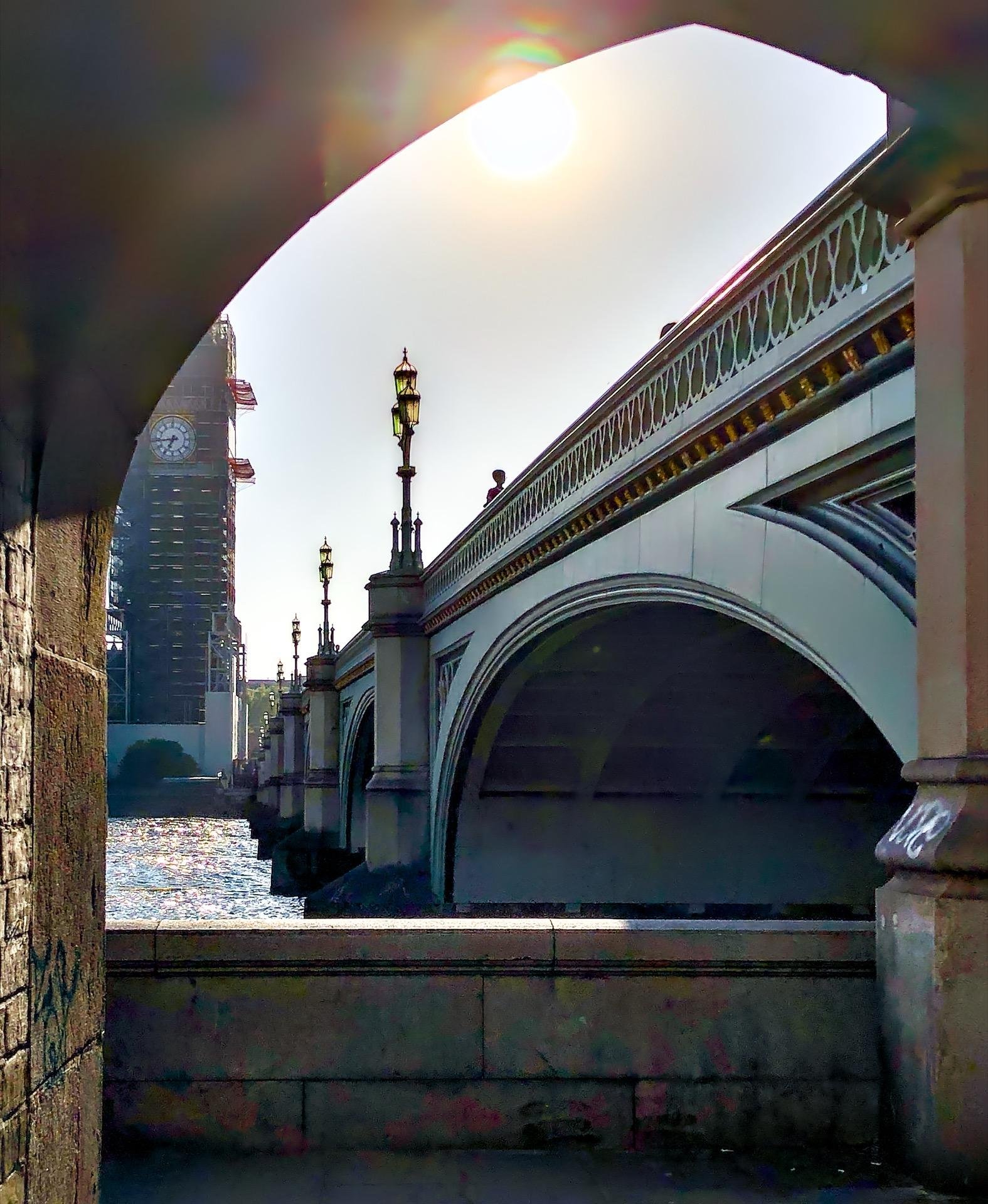 Arched bridge with ornate street lamps and railings viewed through a dark stone archway. The bridge spans over a shimmering body of water, and sunlight creates a flare in the upper left corner. A clock tower partially obscured by the bridge looms in the background.