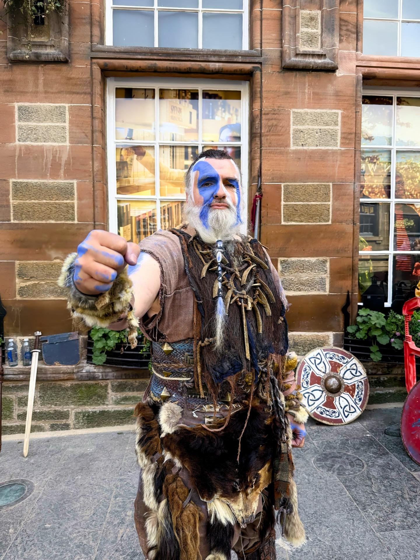 A man in traditional Scottish tribal costume extending his hand toward the camera. He has a blue and white painted face, wearing a fur and leather outfit with ornate accessories, including feathers and metal chains. Behind him is a stone building with a large window reflecting the street scene. A decorative shield rests against the building beside him.