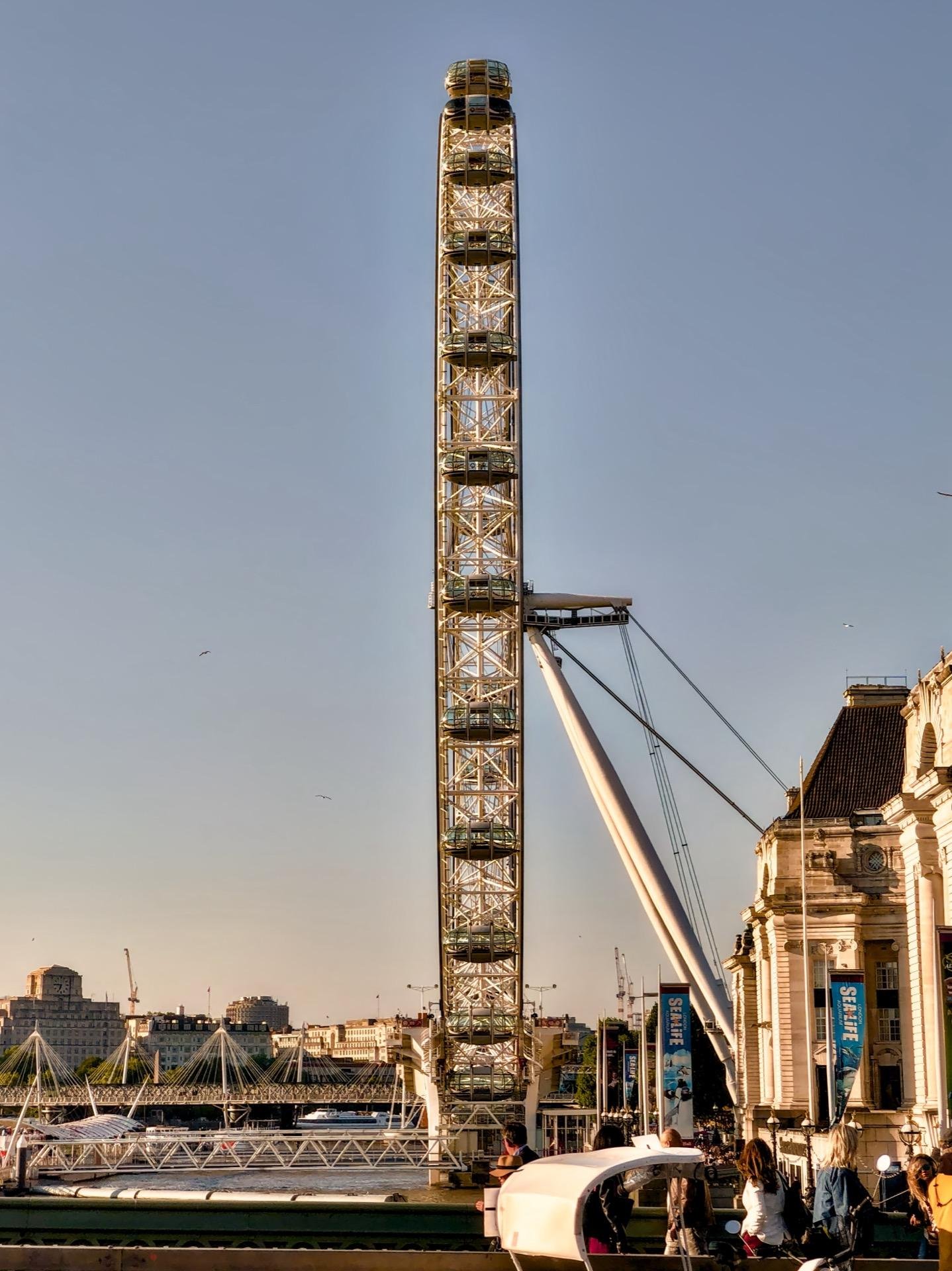 Large Ferris wheel with glass-enclosed capsules against a clear blue sky, with a suspension bridge cable on the right and architectural details of a historic building visible to the right. Below, people are seen walking and sitting near a car with an open trunk, suggesting a lively urban scene.