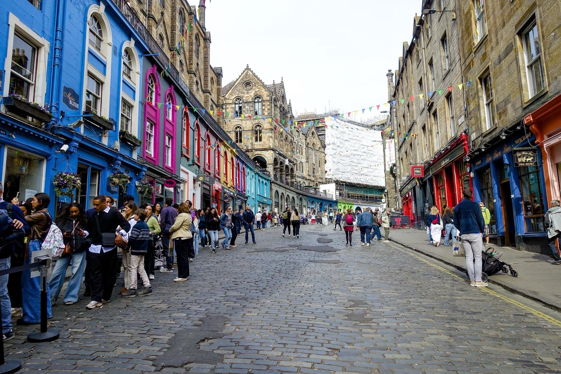 Cobblestone street filled with people, flanked by vibrant blue and red buildings, leading to an old gothic-style structure. A row of people stands to the left, queuing outside a blue building. Multicolored flags are strung above the street.