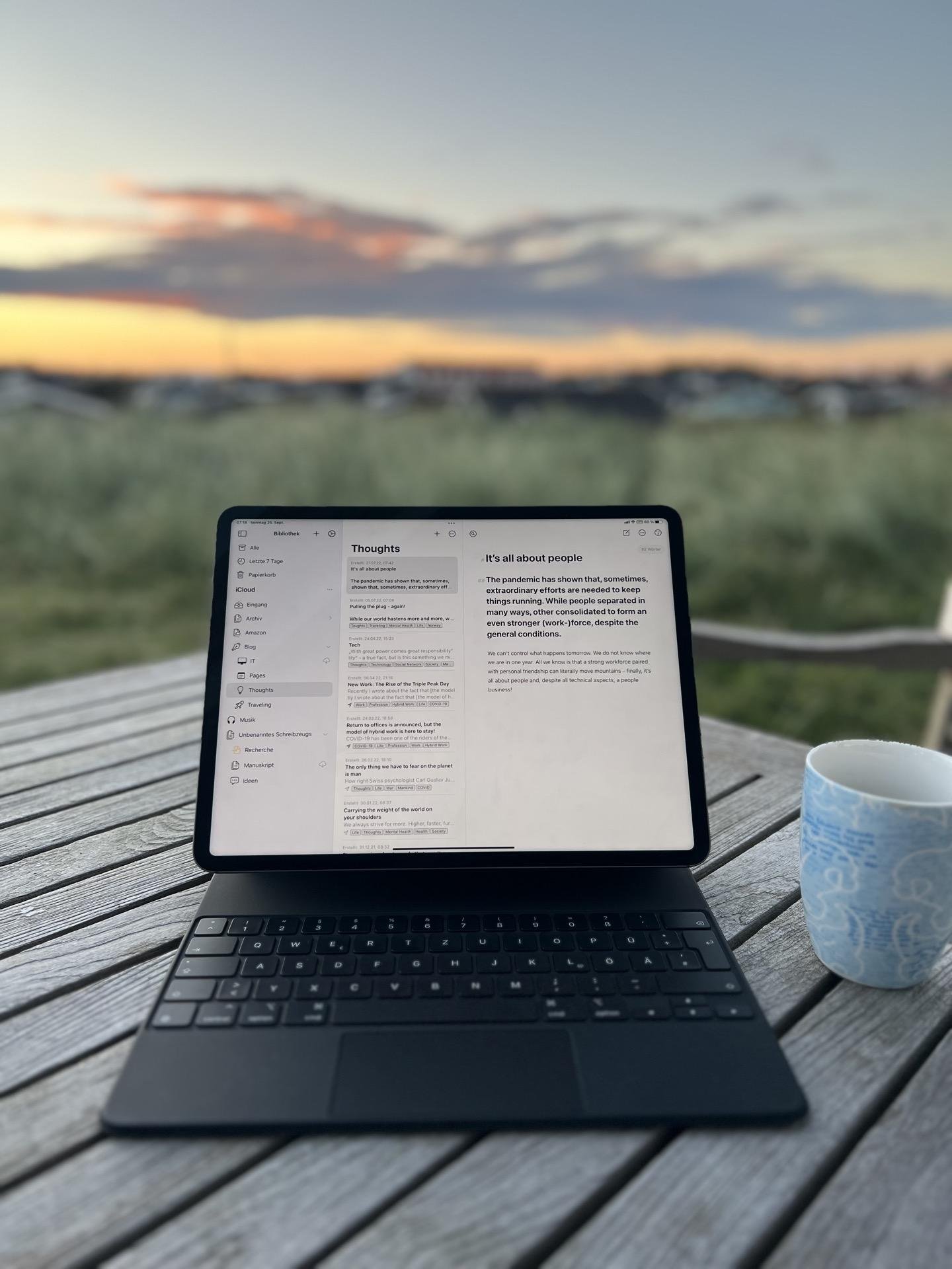 Tablet with keyboard on a wooden table displaying a text document titled "Thoughts" with various bullet points about people and pandemic effects. Beside the tablet is a blue patterned mug. In the background, a blurred landscape features a sunset sky with streaks of orange and blue clouds.