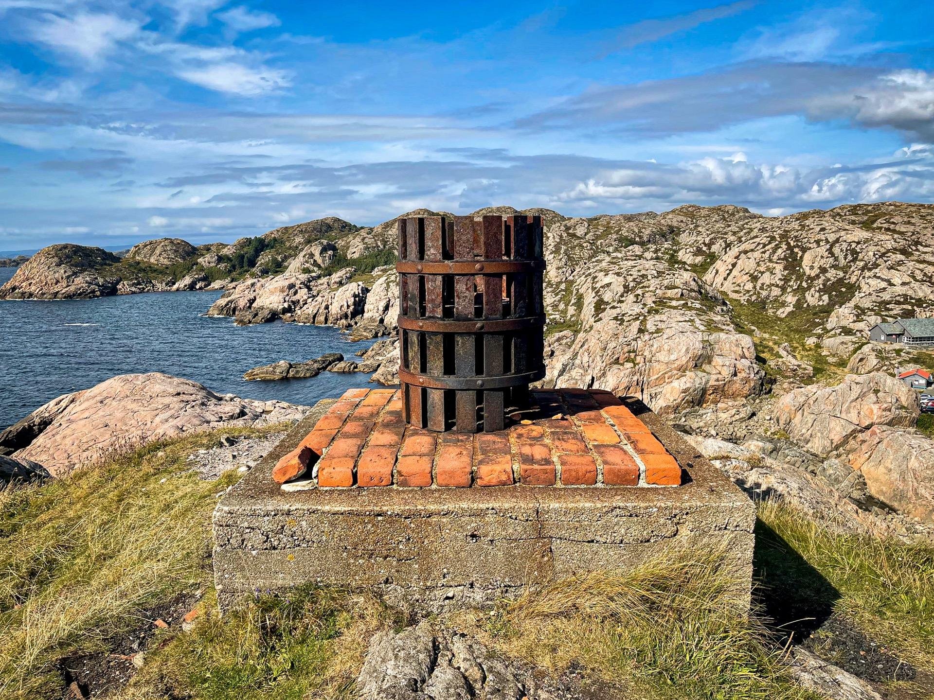 Rusty circular metal object mounted on a brick platform overlooking a rocky coastline with scattered greenery under a blue sky with clouds. In the background, rocky hills and water visible.