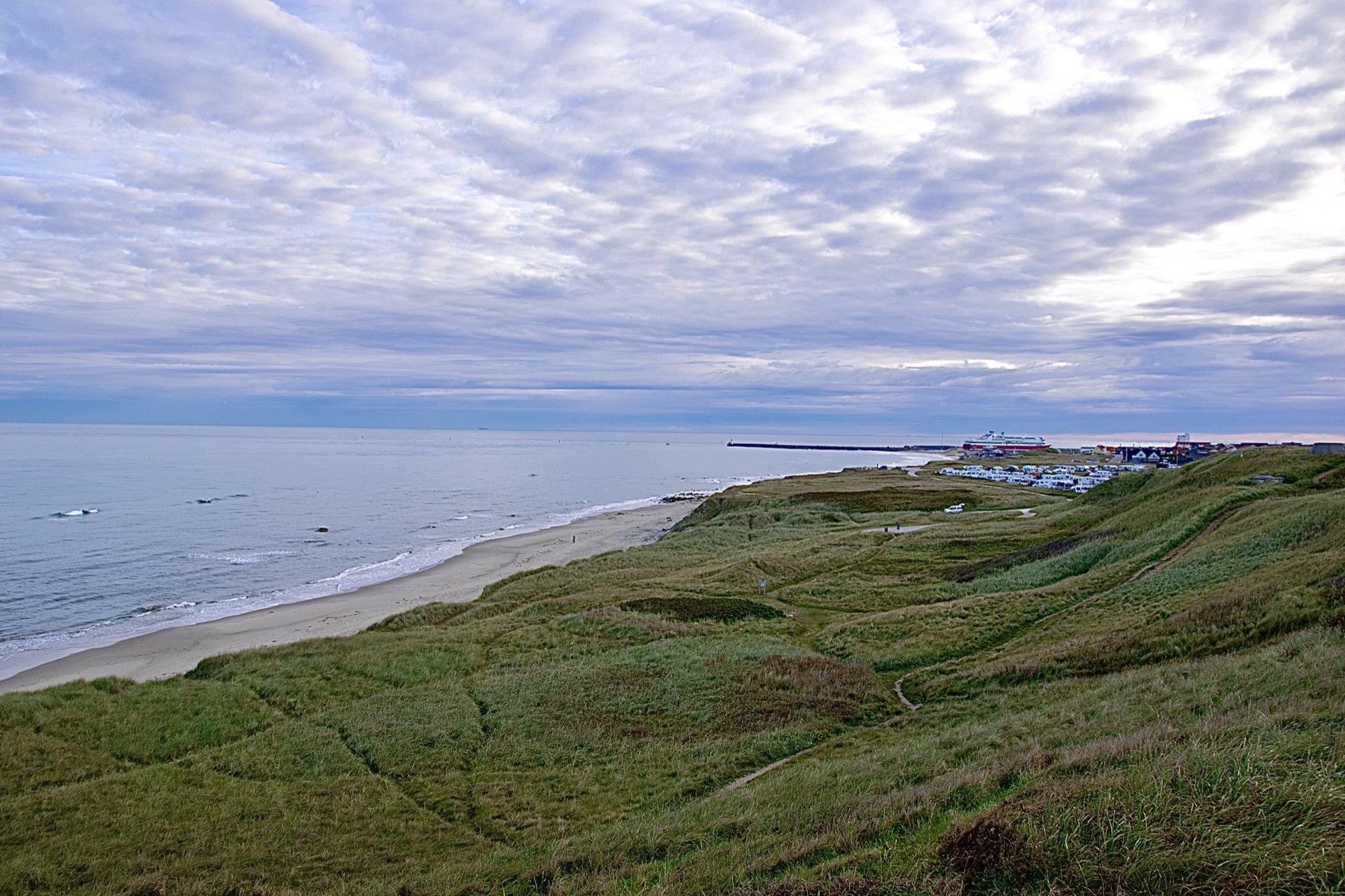 Coastal landscape featuring undulating sand dunes covered with lush green grass, overlooking a calm sea. A cloudy sky blankets the scene, punctuated by diffused sunlight. In the distance, a small town with buildings including what appears to be a large ship-like structure is visible along the shoreline. Paths weave through the dunes, suggesting frequent foot traffic or maintenance.