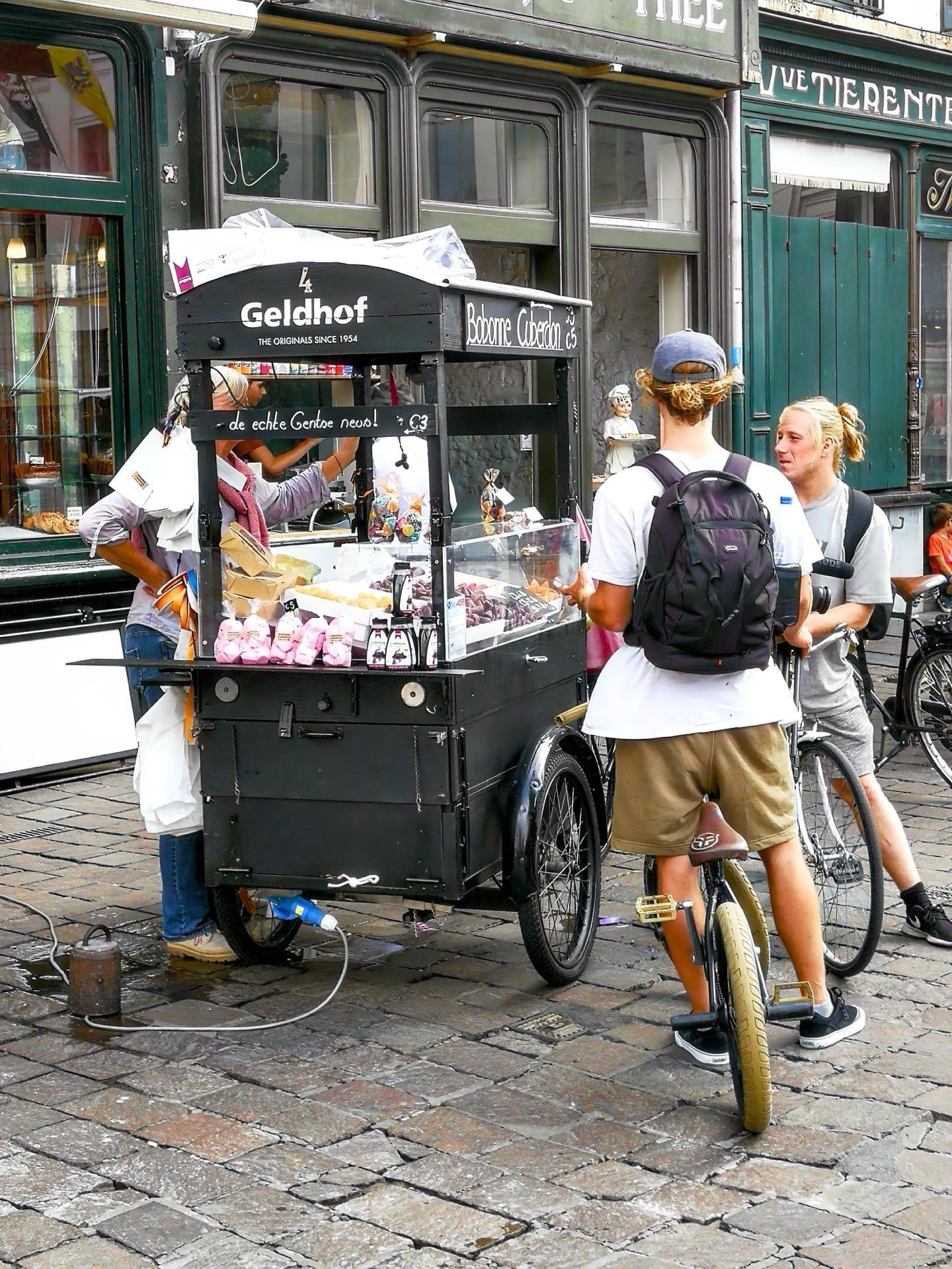 Two men converse near a mobile candy cart on a cobblestone street. The cart, labeled "Geldhof The Original Since 1964," displays various confections. One man stands with a backpack, facing the vendor who multitasks handling the goods.