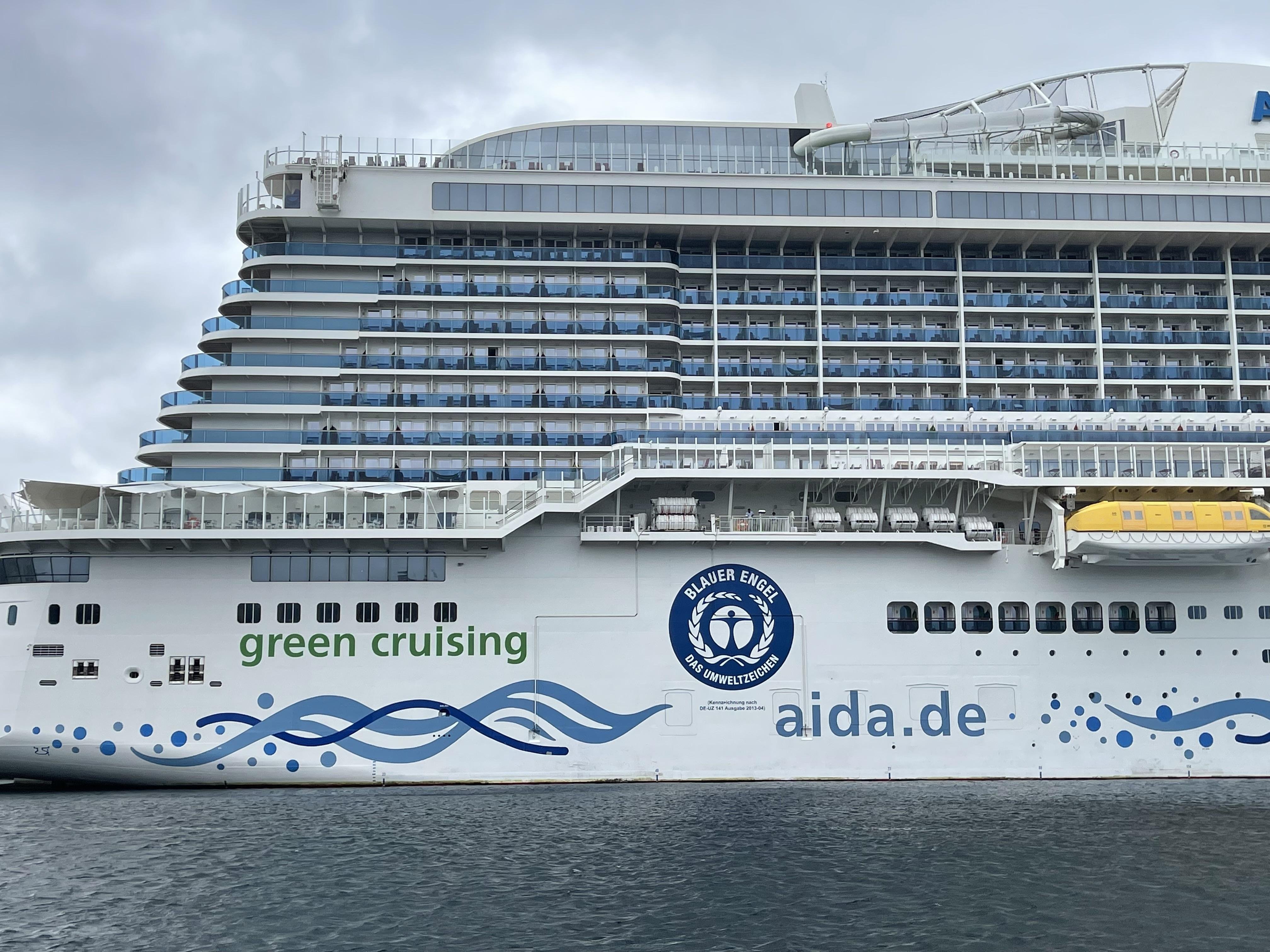 Side view of a large white cruise ship with multiple decks featuring rows of balconies. The hull displays the text "green cruising" and the logo of AIDA Cruises, along with a blue graphic design of waves. A yellow lifeboat hangs on the side amidst cloudy skies and calm sea in the background.
