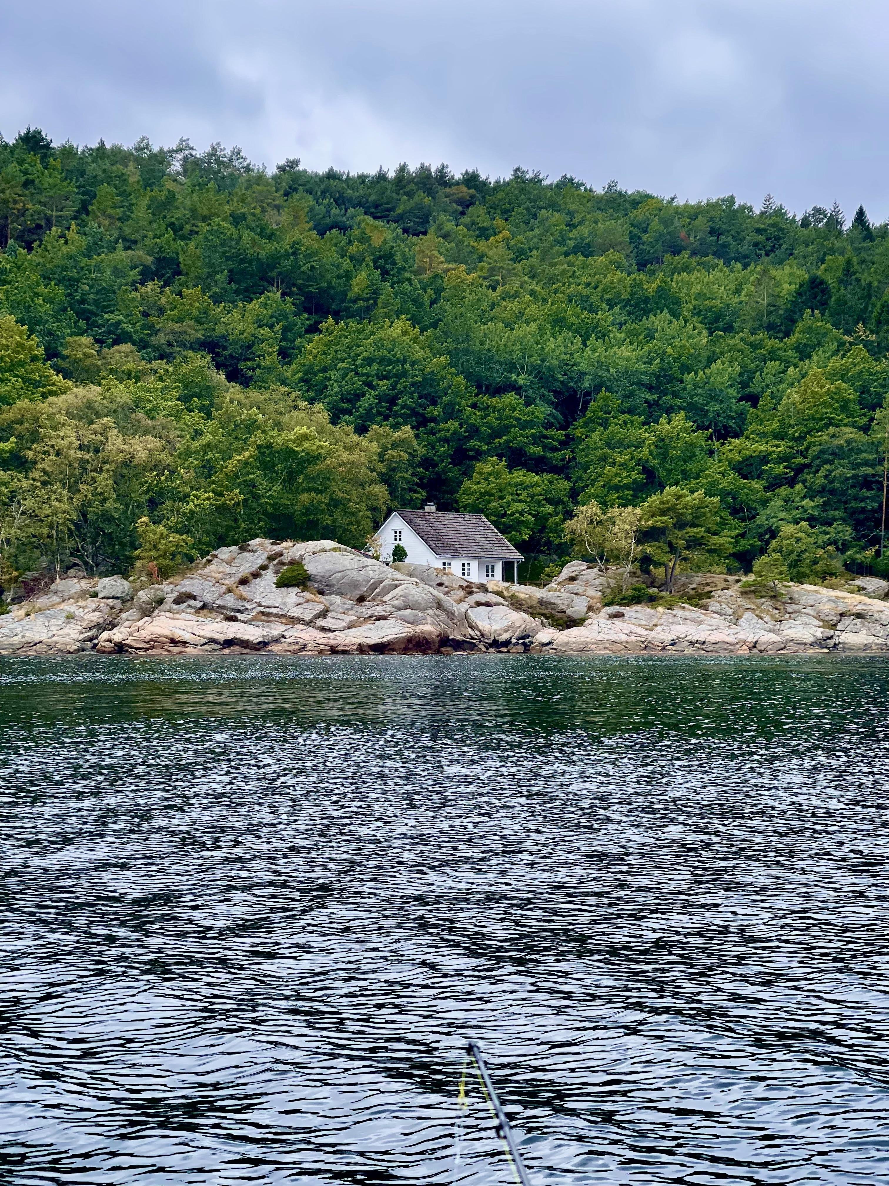A small white house with a pitched roof sits nestled among rocks at the edge of a dense, green forest. In the foreground, rippled dark blue water extends towards the viewer, with a hint of a boat railing visible at the bottom of the frame. Overcast skies contribute to the moody atmosphere of the scene.
