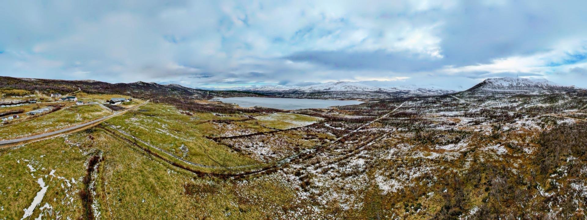 Panoramic winter landscape featuring sparsely snow-covered hills with patches of barren vegetation and a network of roads. In the background, a calm lake nestles among the hills under a cloudy sky.