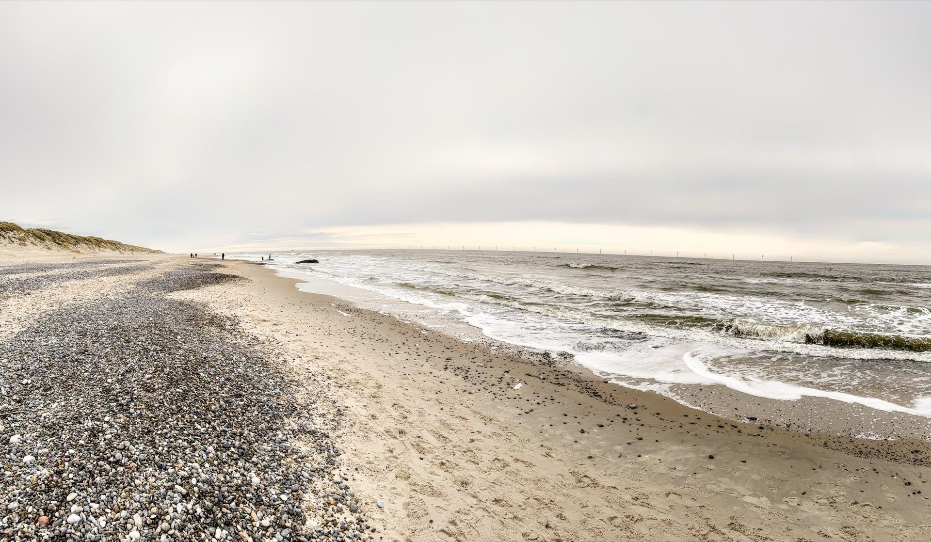 Panoramic view of a pebble-strewn beach leading into a sandy shore beside calm sea waves, under an overcast sky with faint figures in the distance and an array of offshore wind turbines visible on the horizon.